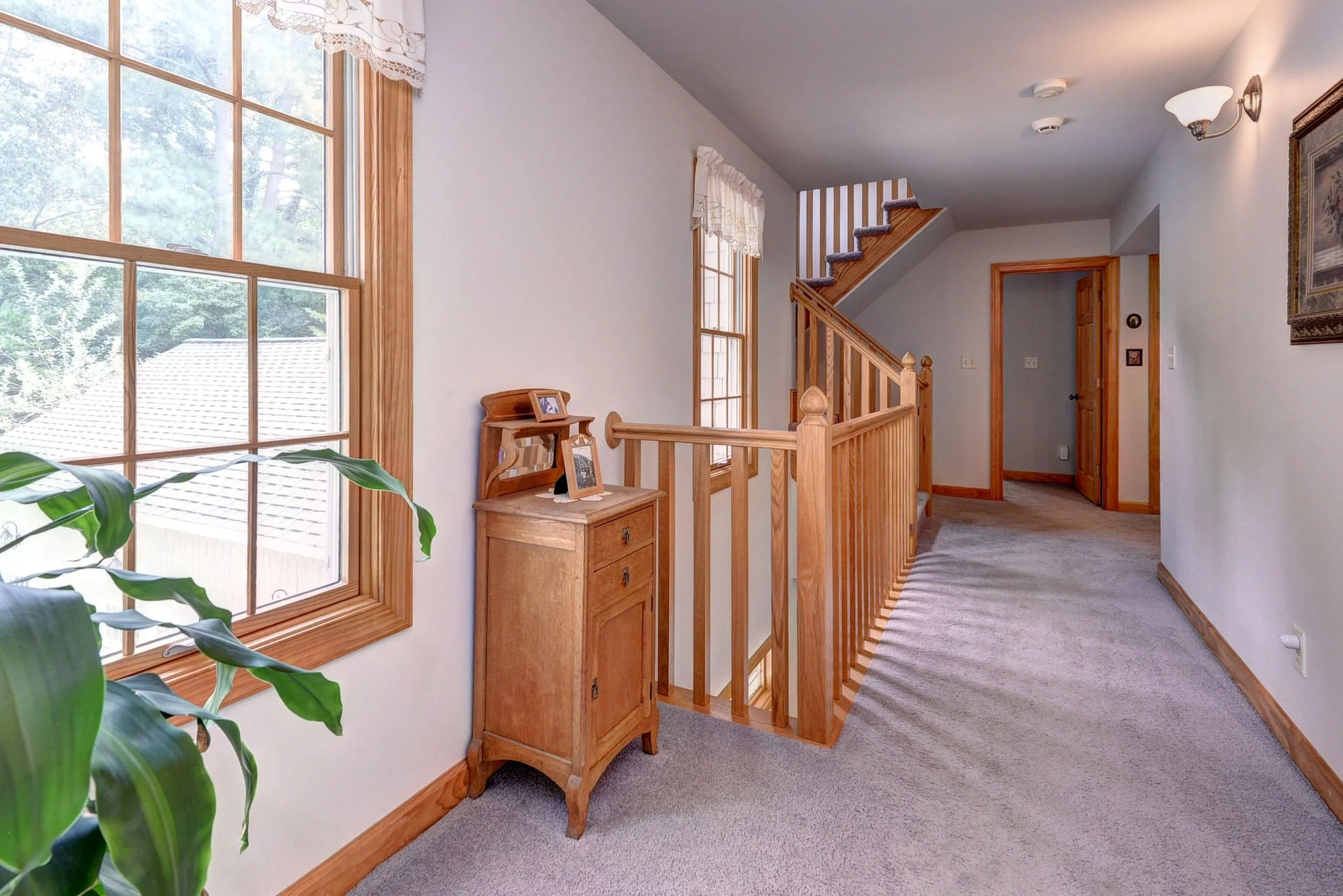 Hallway with large windows, a wooden staircase, and a small wooden cabinet with framed photos.
