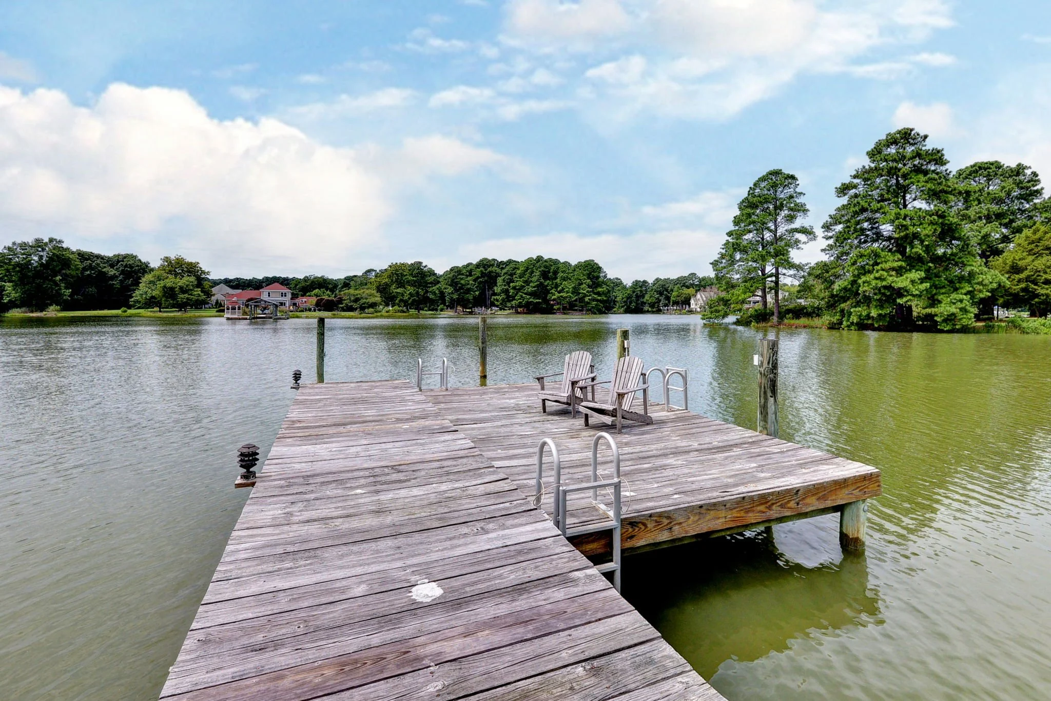 A wooden dock extending into a calm lake with two adirondack chairs, surrounded by trees and a few houses in the distance under a partly cloudy sky.