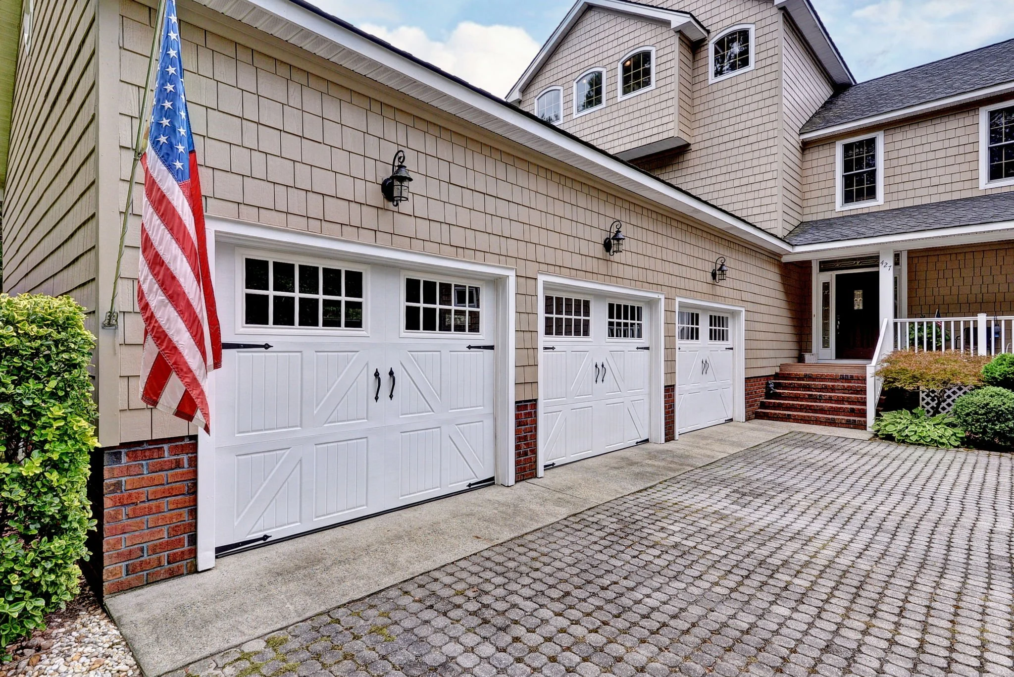 Three white garage doors with windows at the top, part of a beige house with shingle siding, an American flag on the left side, and a concrete driveway. There are outdoor lights above each garage door and steps leading to the front door on the right.