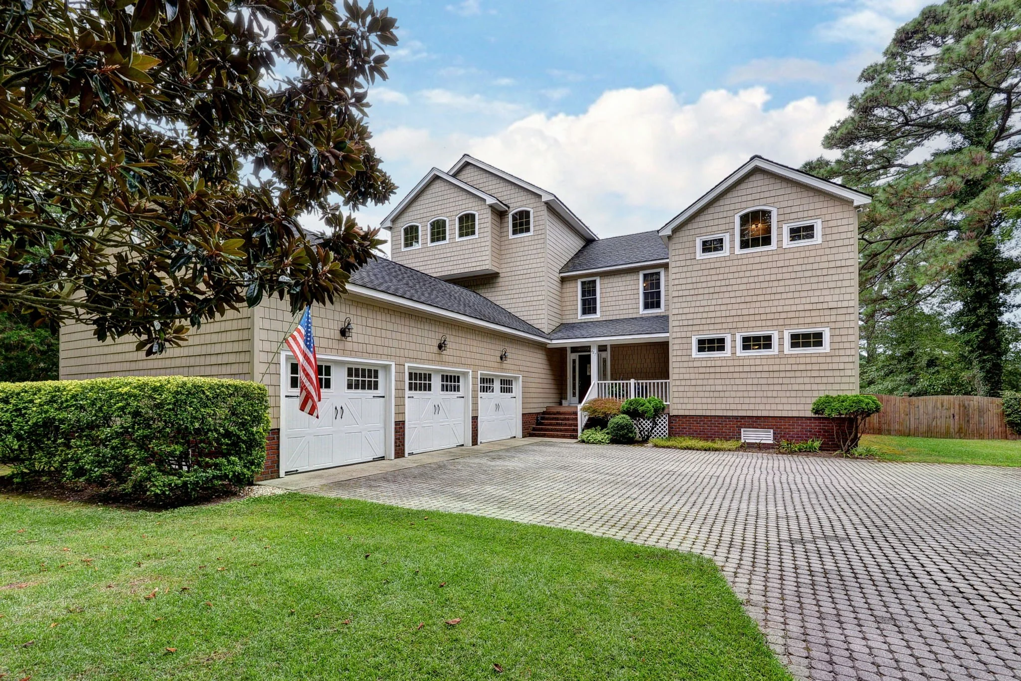 Large suburban house with a brick and shingle exterior, attached garage, landscaped yard, and American flag displayed near the garage, under a partly cloudy sky.