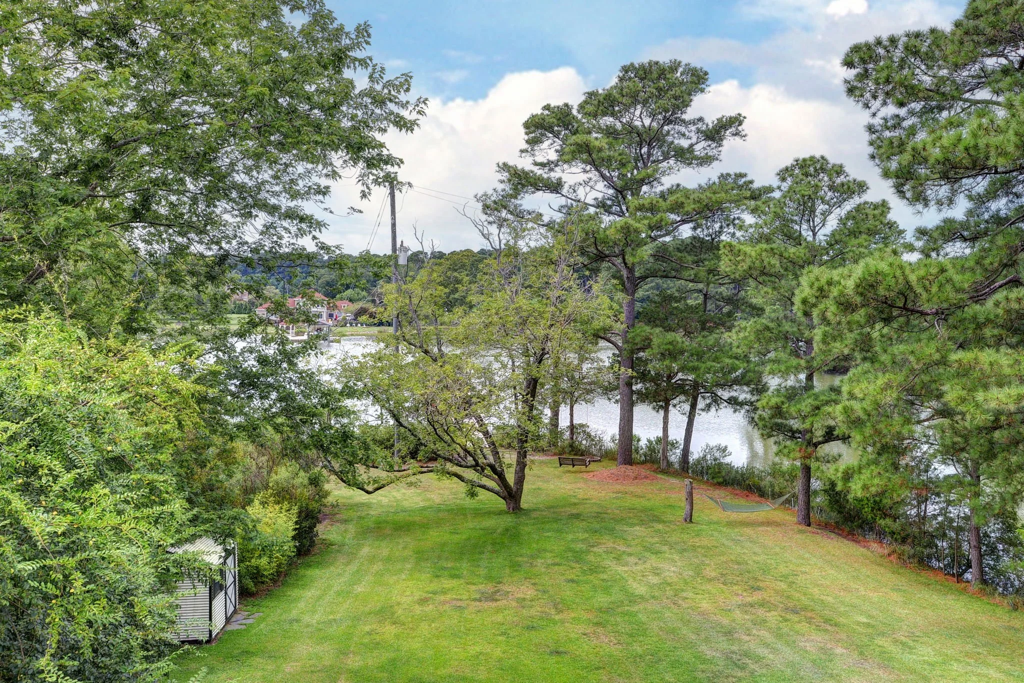 A grassy backyard with trees near a lake. There are benches, a hammock, and a shed visible.