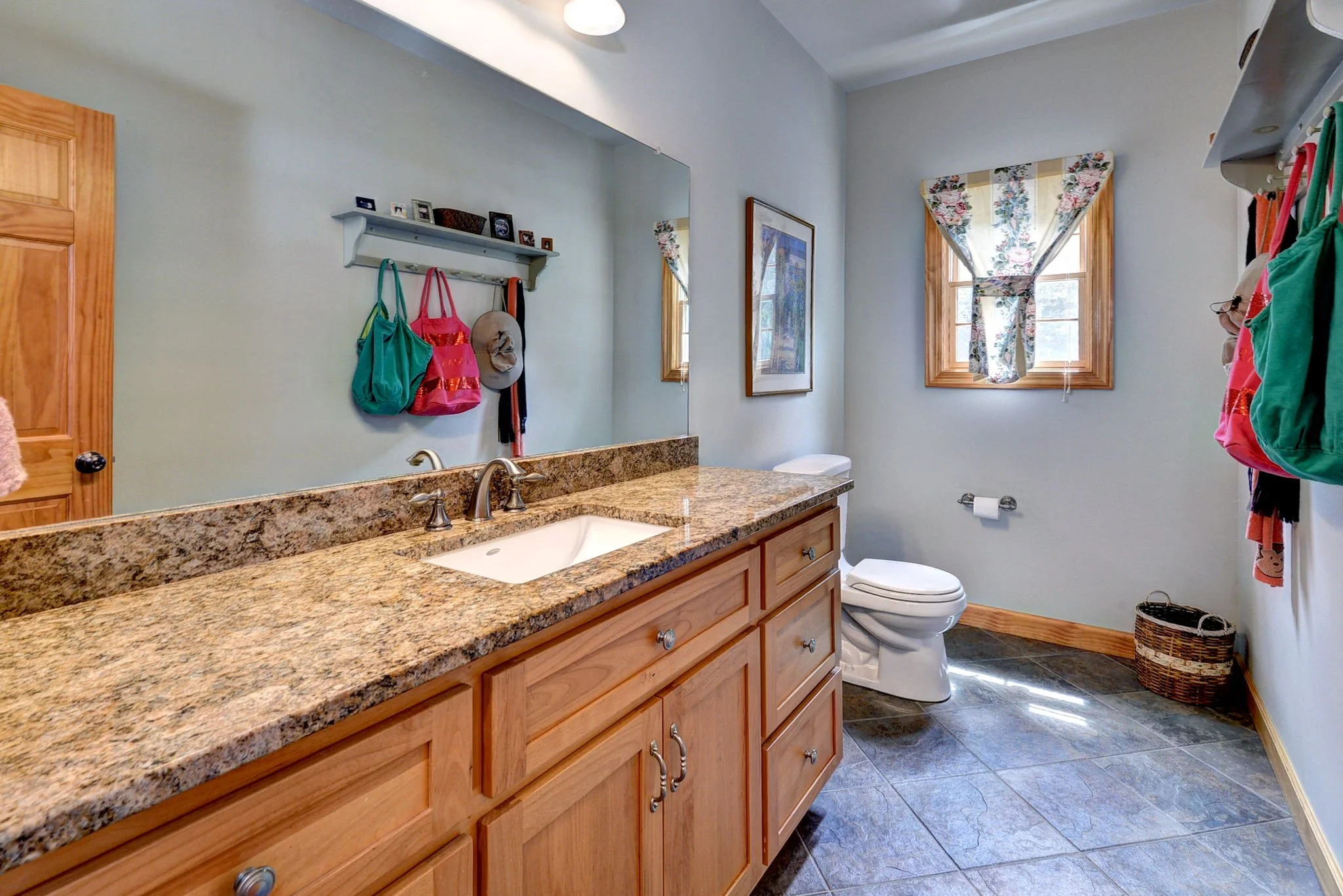 Bathroom with wooden vanity, granite countertop, white sink, bronze fixtures, mirror, two small windows with floral curtains, toilet, wall shelf with hanging bags and hats, framed art, wicker basket, tiled floor.