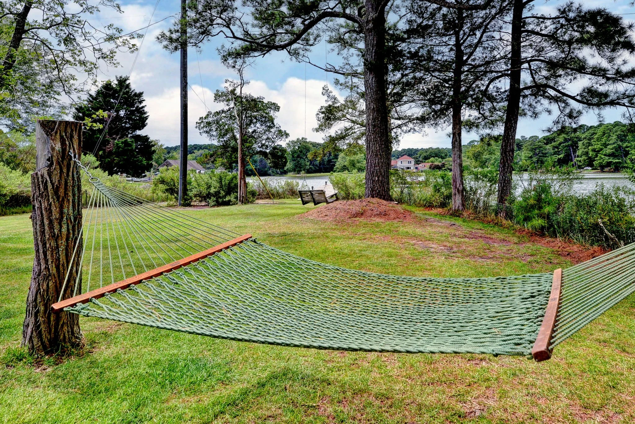 Green hammock tied between two trees near a lake with chairs in the background.