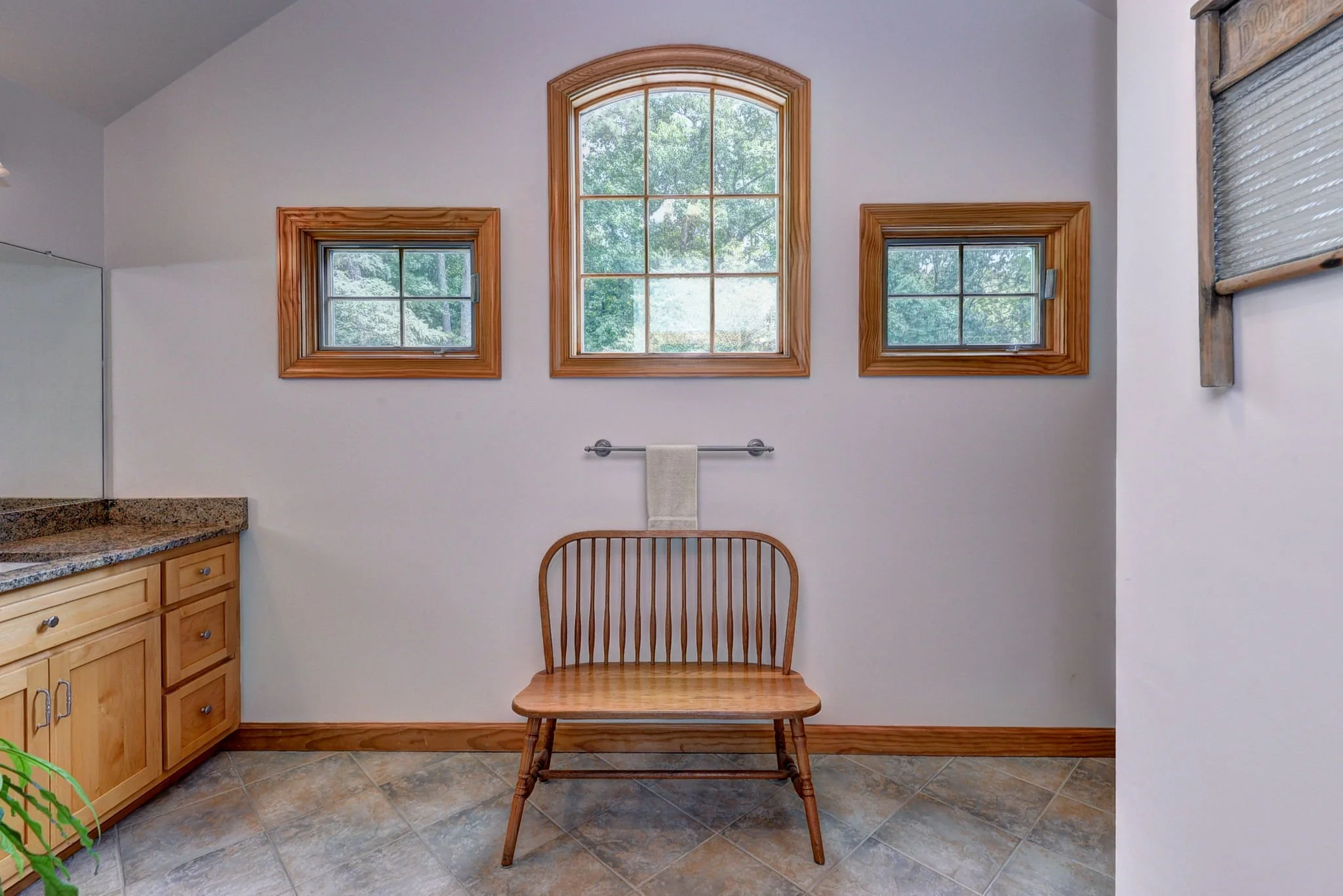 A bathroom with three windows, a wooden bench, and a towel rack with a towel, in front of a white wall and a granite countertop.