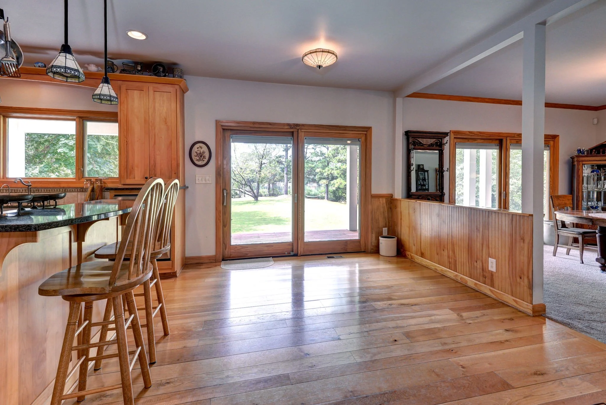 Interior view of a house showing a wooden floor, sliding glass doors leading outside, a kitchen counter with bar stools, and a partial view of a dining room with a wooden cabinet and a dining table.