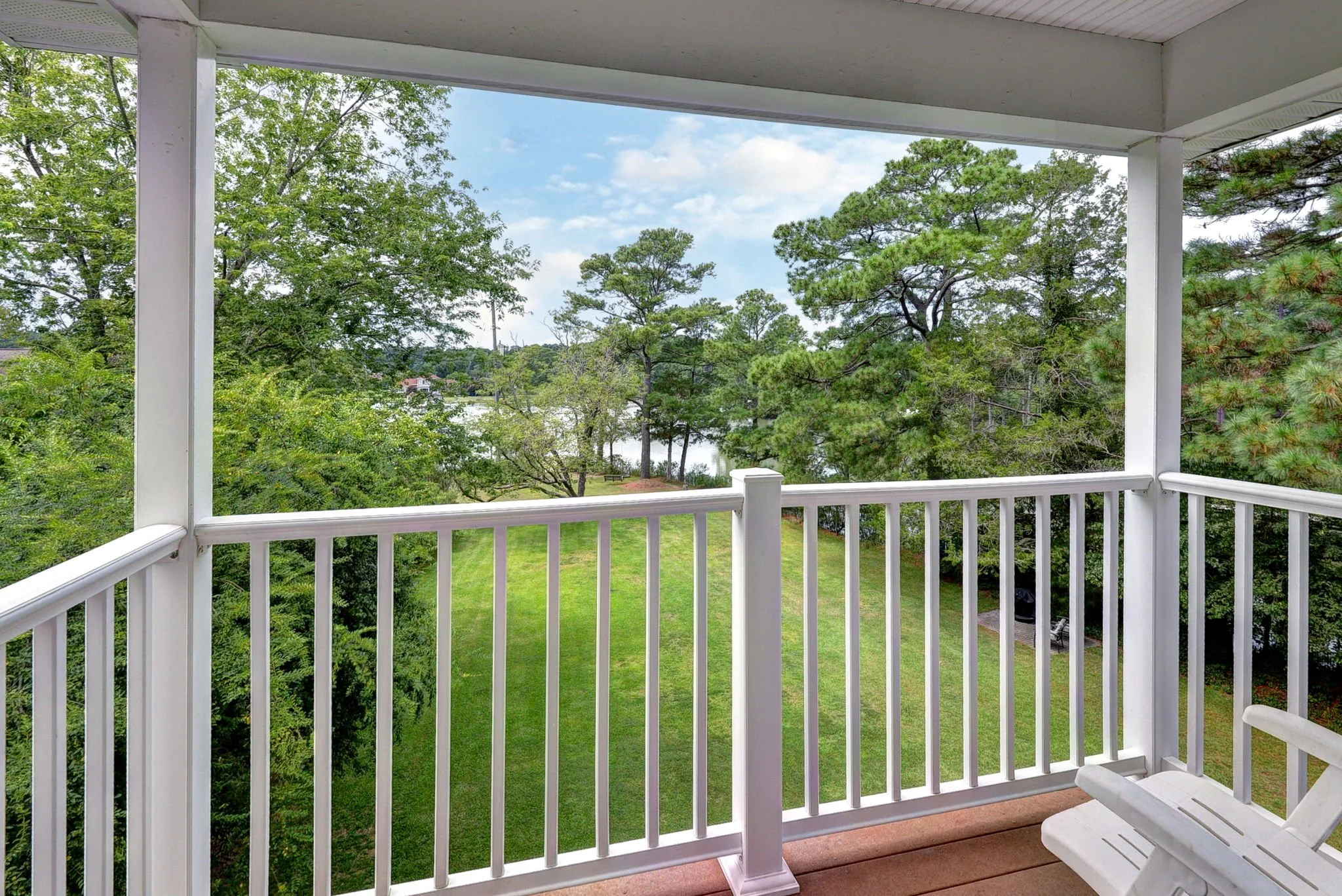 View from a balcony overlooking a lush green yard, trees, and a body of water in the distance, with a partly cloudy sky.