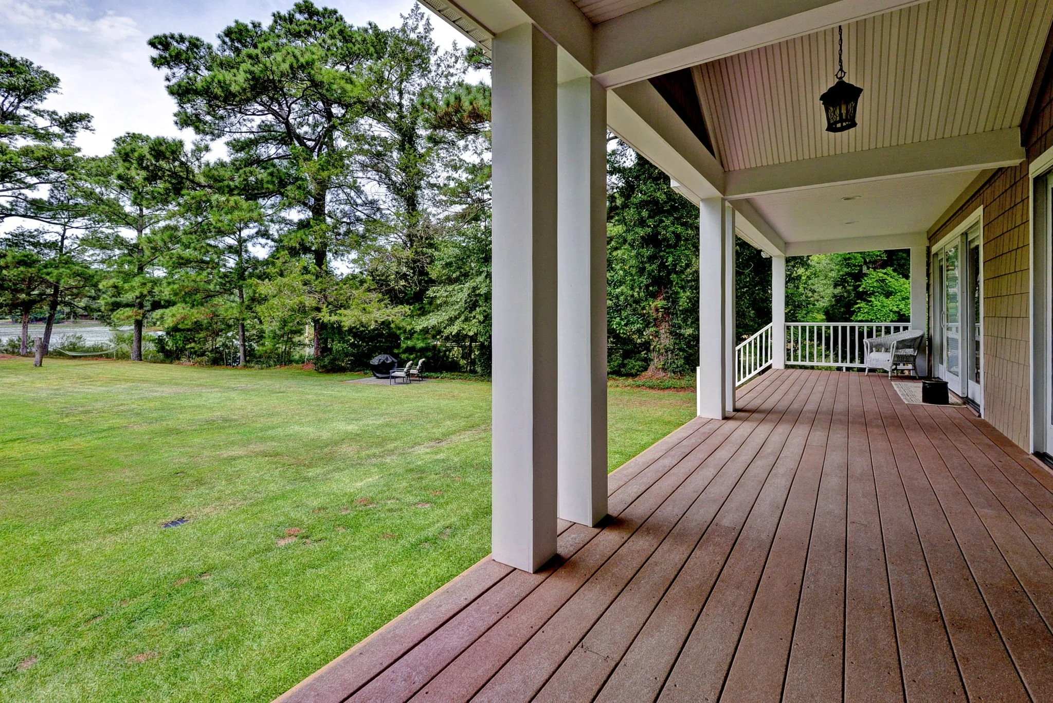 Covered wooden porch overlooking a grassy yard with trees and outdoor furniture, including a grill and chairs.