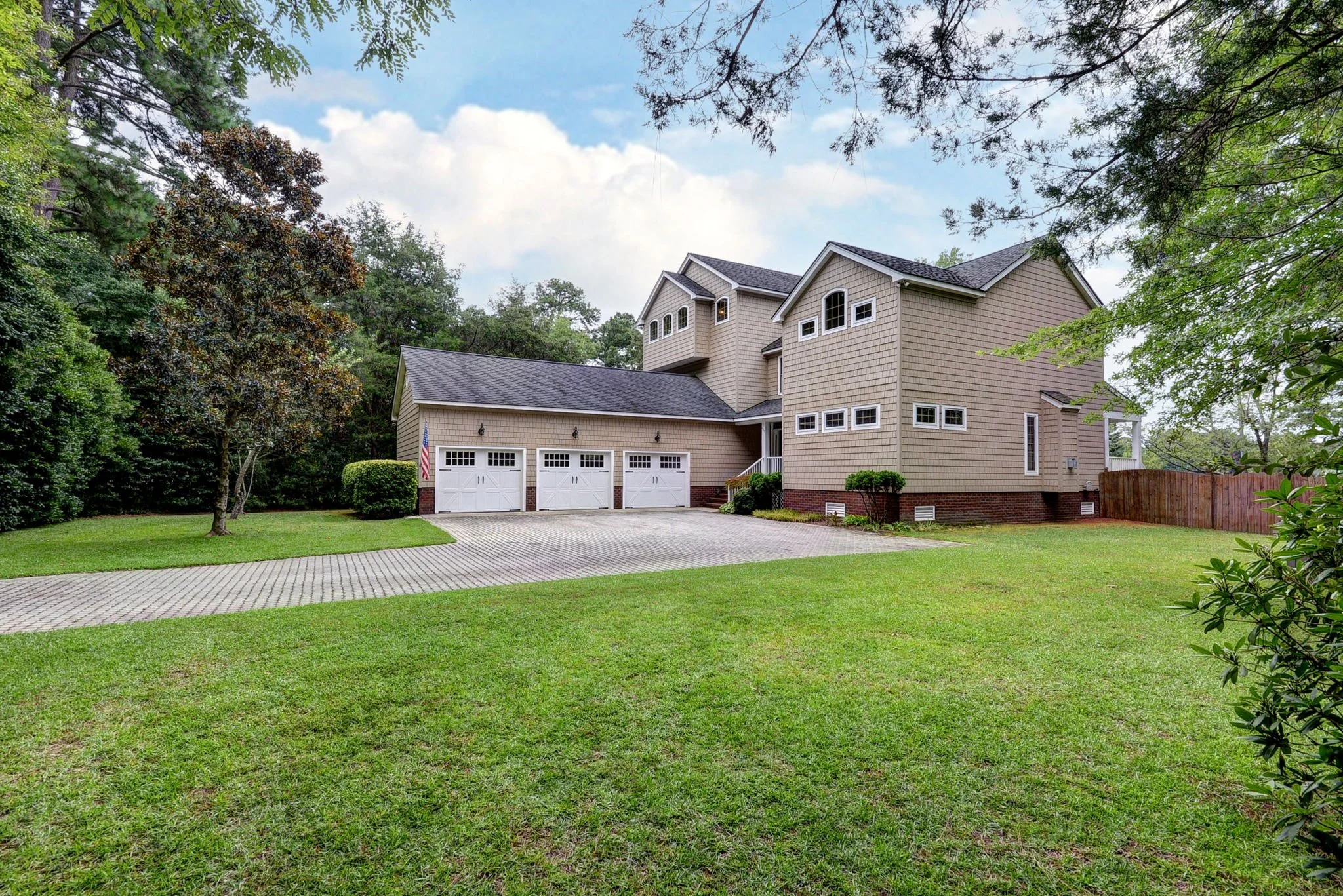 Large beige house with attached three-car garage, surrounded by green lawn and trees, under a partly cloudy sky.