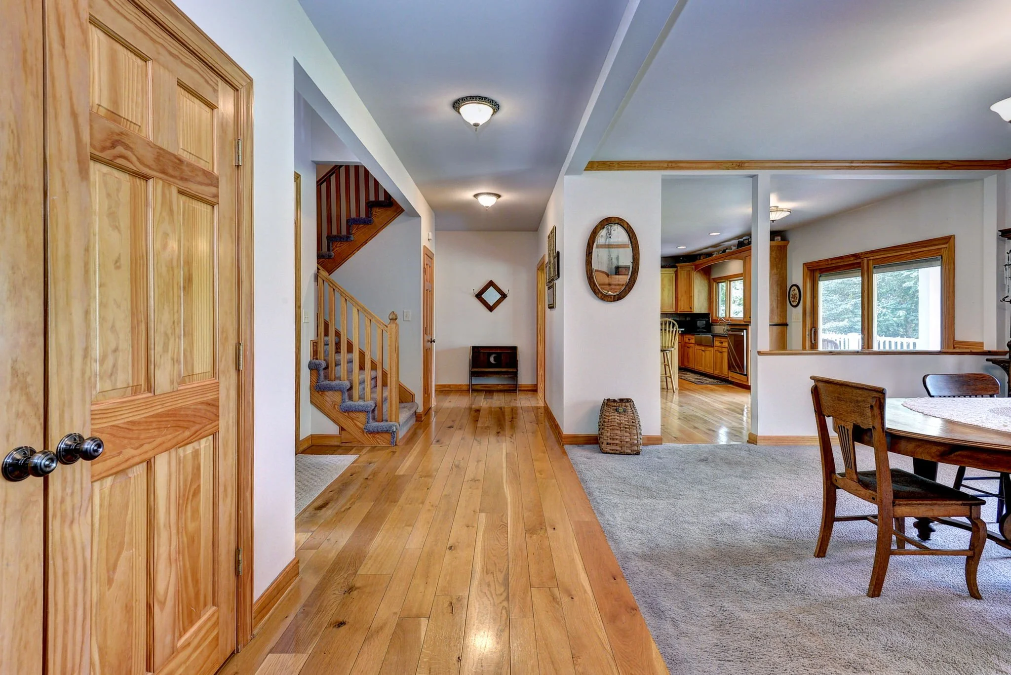 Interior view of a house with wooden floors, staircase with blue carpet, and a dining area with chairs around a wooden table. Walls are white with wooden trim, and there is a kitchen with wooden cabinets and large windows.