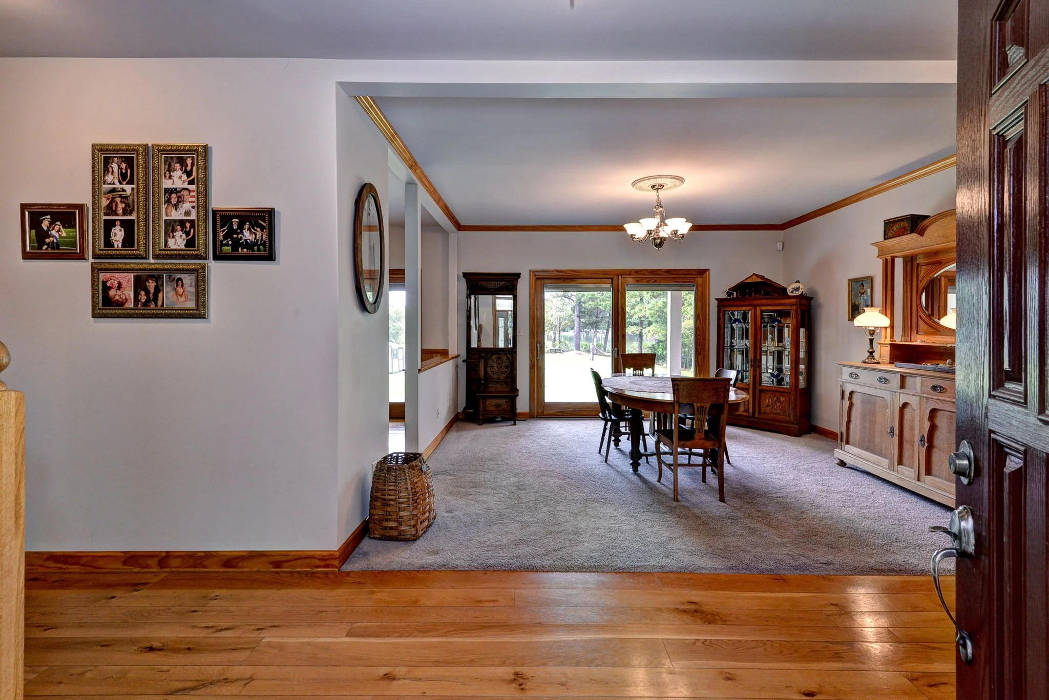 View of a dining room with a wooden table and chairs, a china cabinet, a sideboard with a lamp, and sliding glass doors leading outside. There is a wall with framed photographs and a mirror.