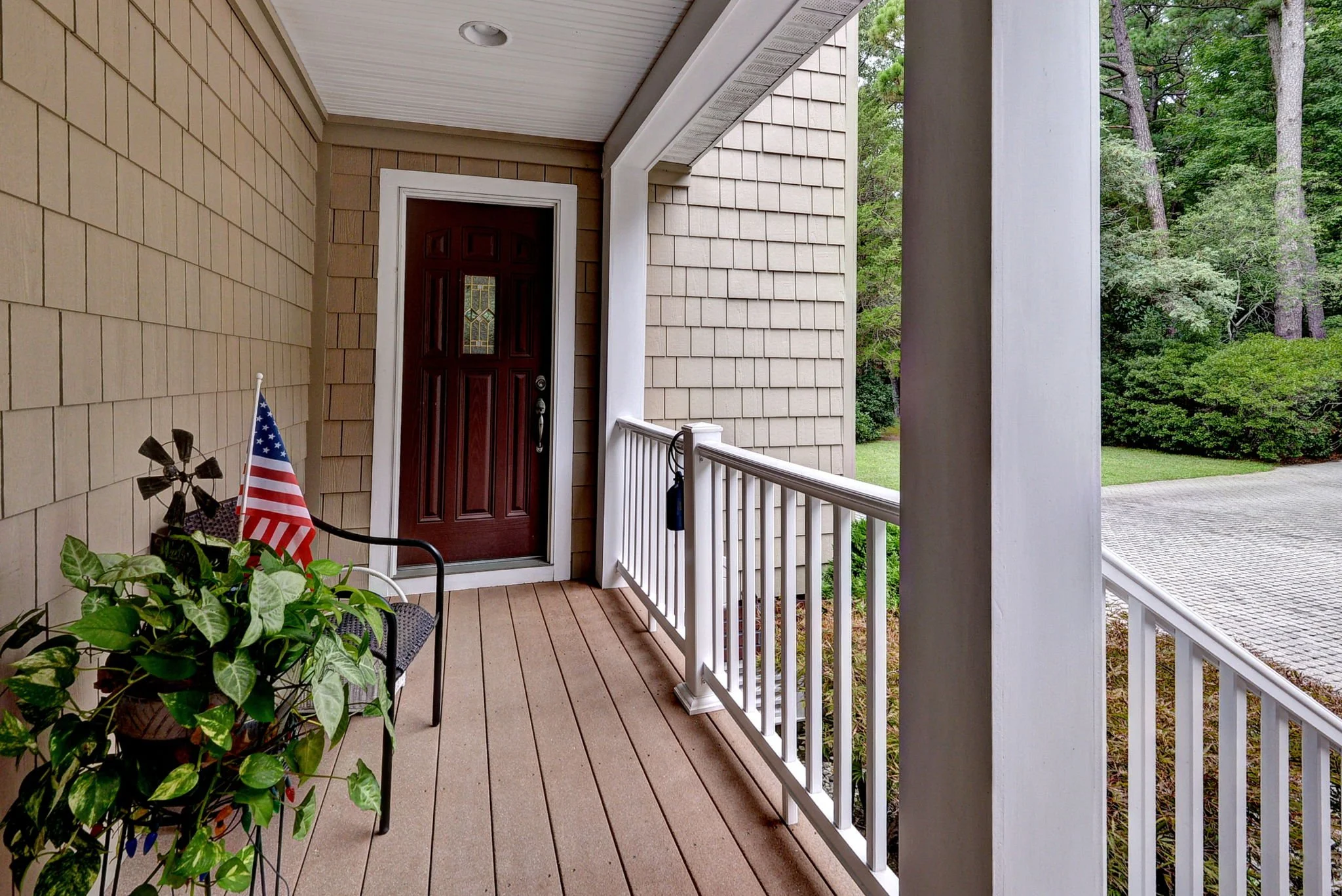 Front porch with a dark wooden door, white railing, potted plant, and American flag, overlooking a driveway and green trees.