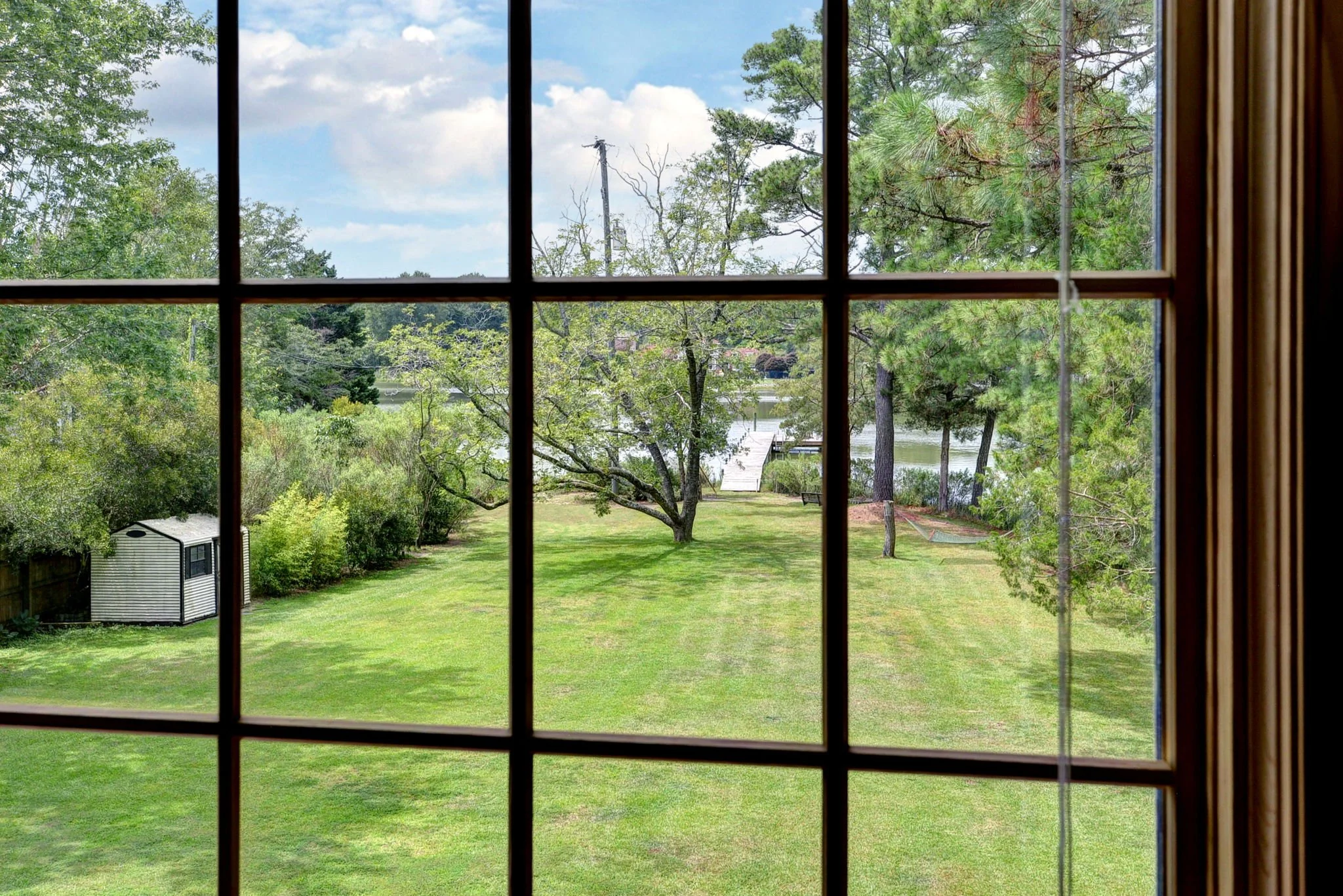 View of a lush green backyard with a tree, a small shed, a dock extending into a lake, and trees surrounding the water outdoors, seen through a window with grid muntins.