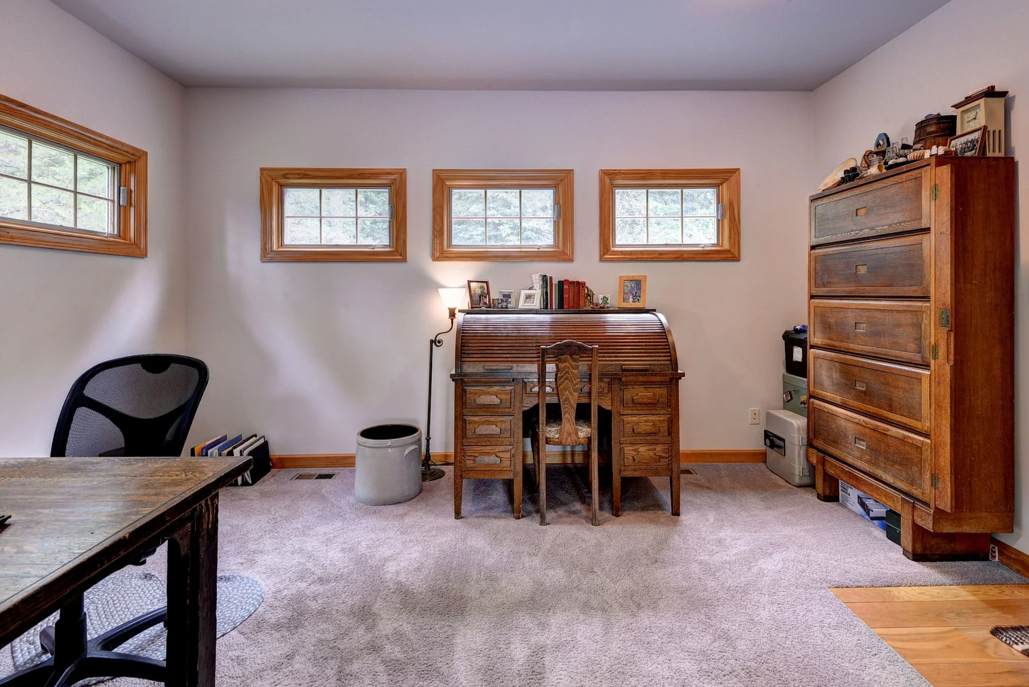 Home office with wooden desk, office chair, and tall wooden dresser against white wall with four small windows.
