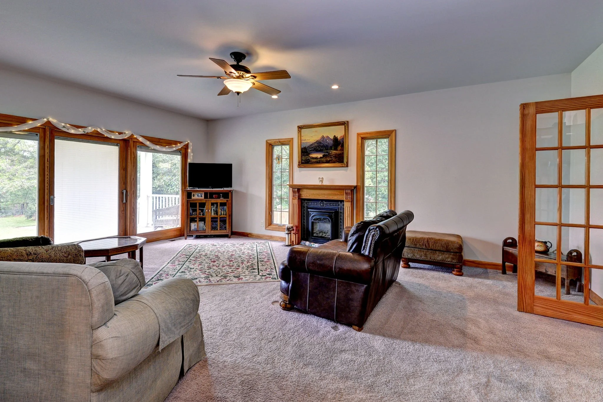 Living room with beige carpet, beige and dark leather couches, a fireplace with a wooden mantle, a small flat-screen TV, windows with wooden frames, and a door leading outside. There are decorative elements like a 'Christmas' ribbon on the window and