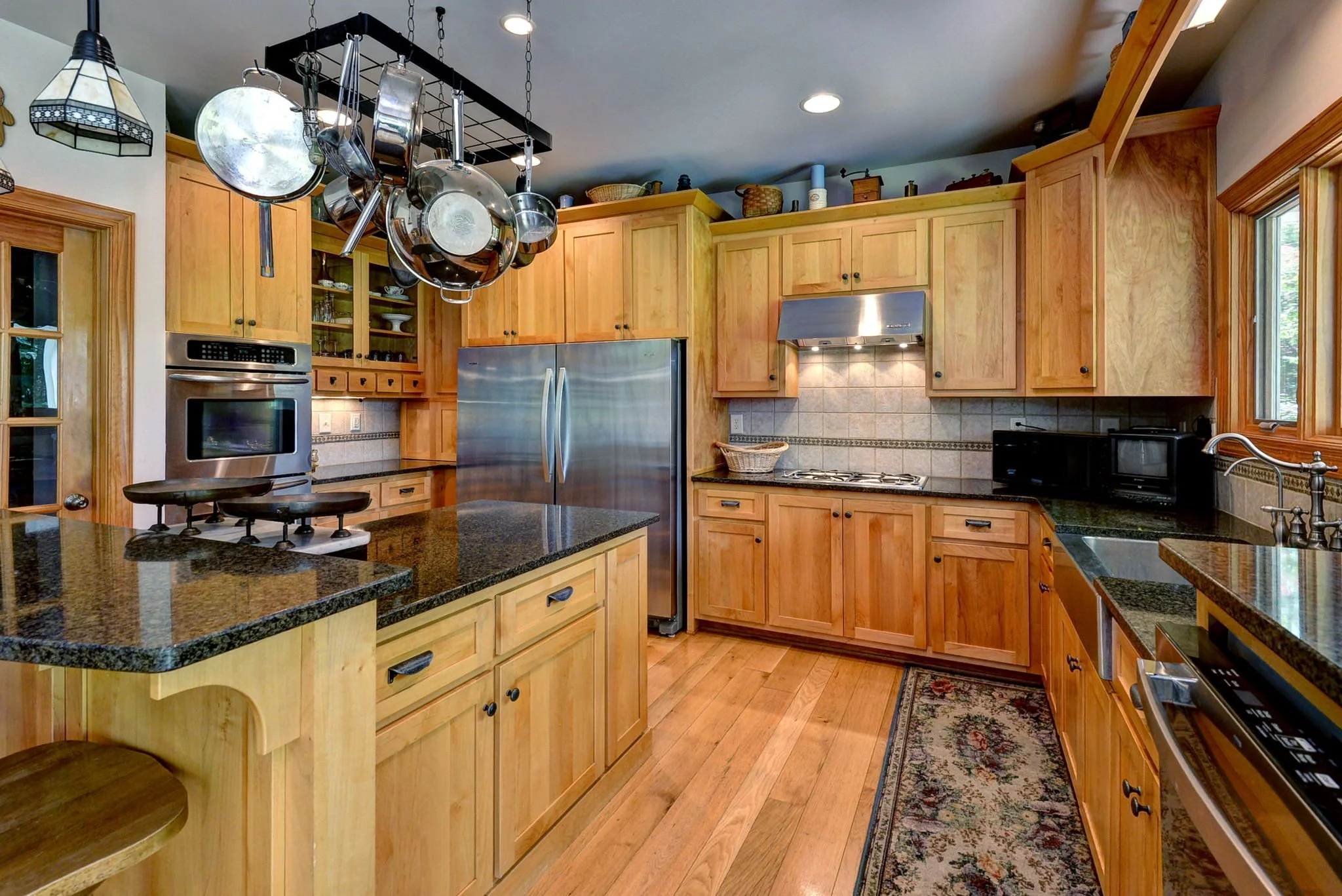 A kitchen with wooden cabinets, a stainless steel refrigerator, and a hanging pot rack with pots and pans. There is a black granite countertop and hardwood floors, with a window above the sink.