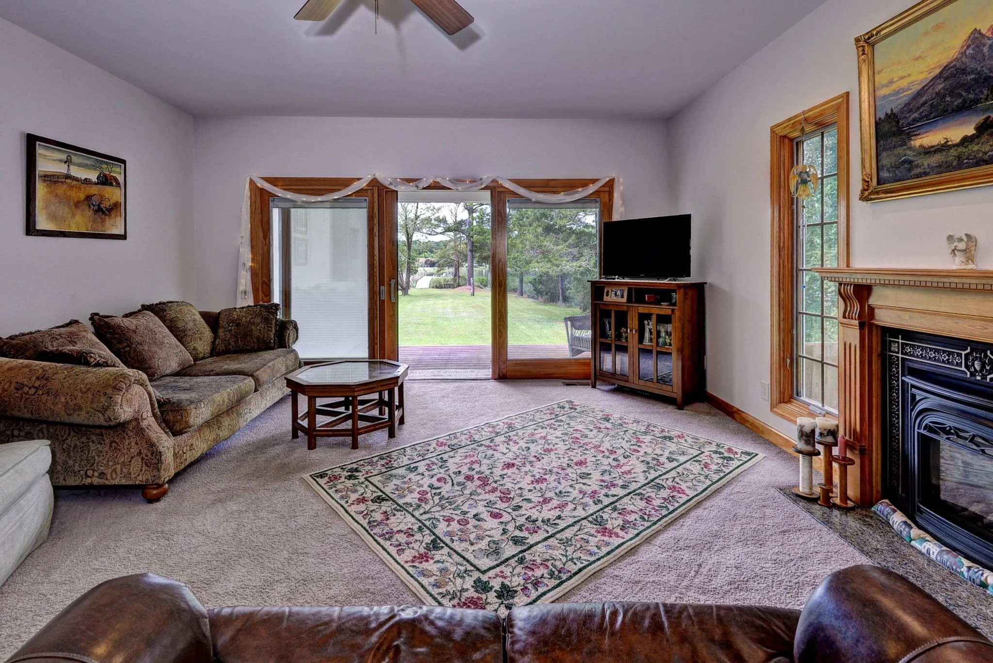 Living room with beige carpet, brown patterned sofa, coffee table, fireplace, flat-screen TV on wooden stand, and large sliding glass door leading to a backyard with grass and trees.