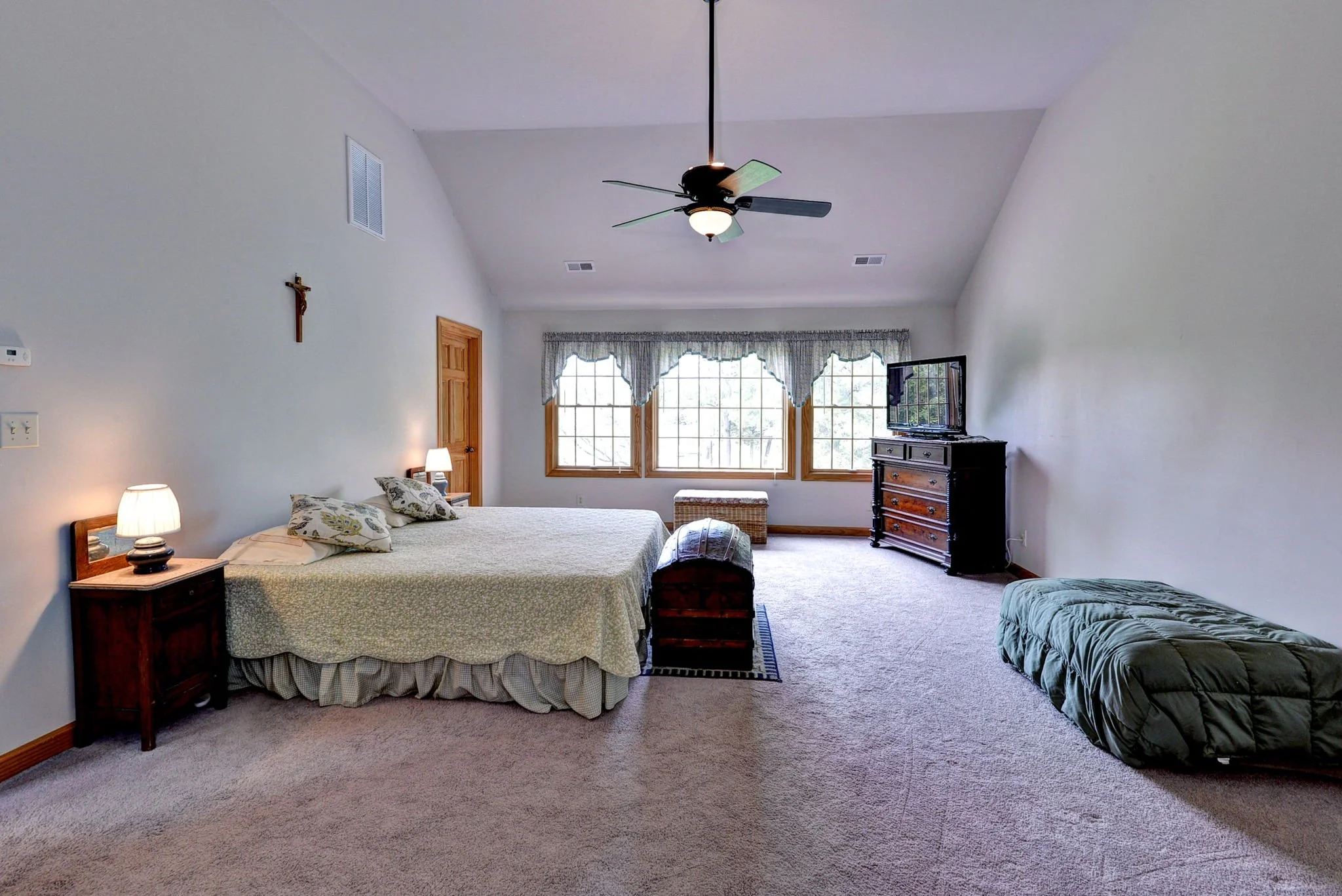 Spacious bedroom with a large bed, nightstands with lamps, windows with curtains, a chest at the foot of the bed, a TV on a dresser, a green floor mattress, and a ceiling fan.