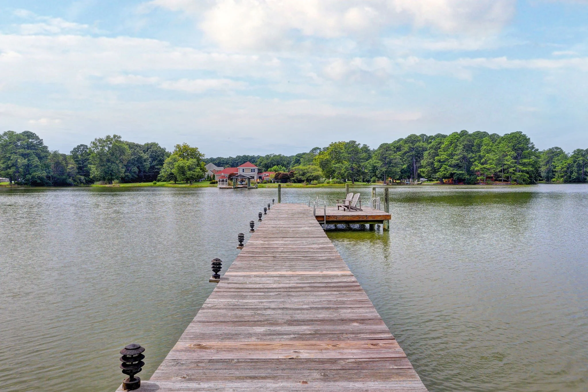 A wooden pier extending over calm lake water with two Adirondack chairs at the end, surrounded by trees and houses in the background on a partly cloudy day.