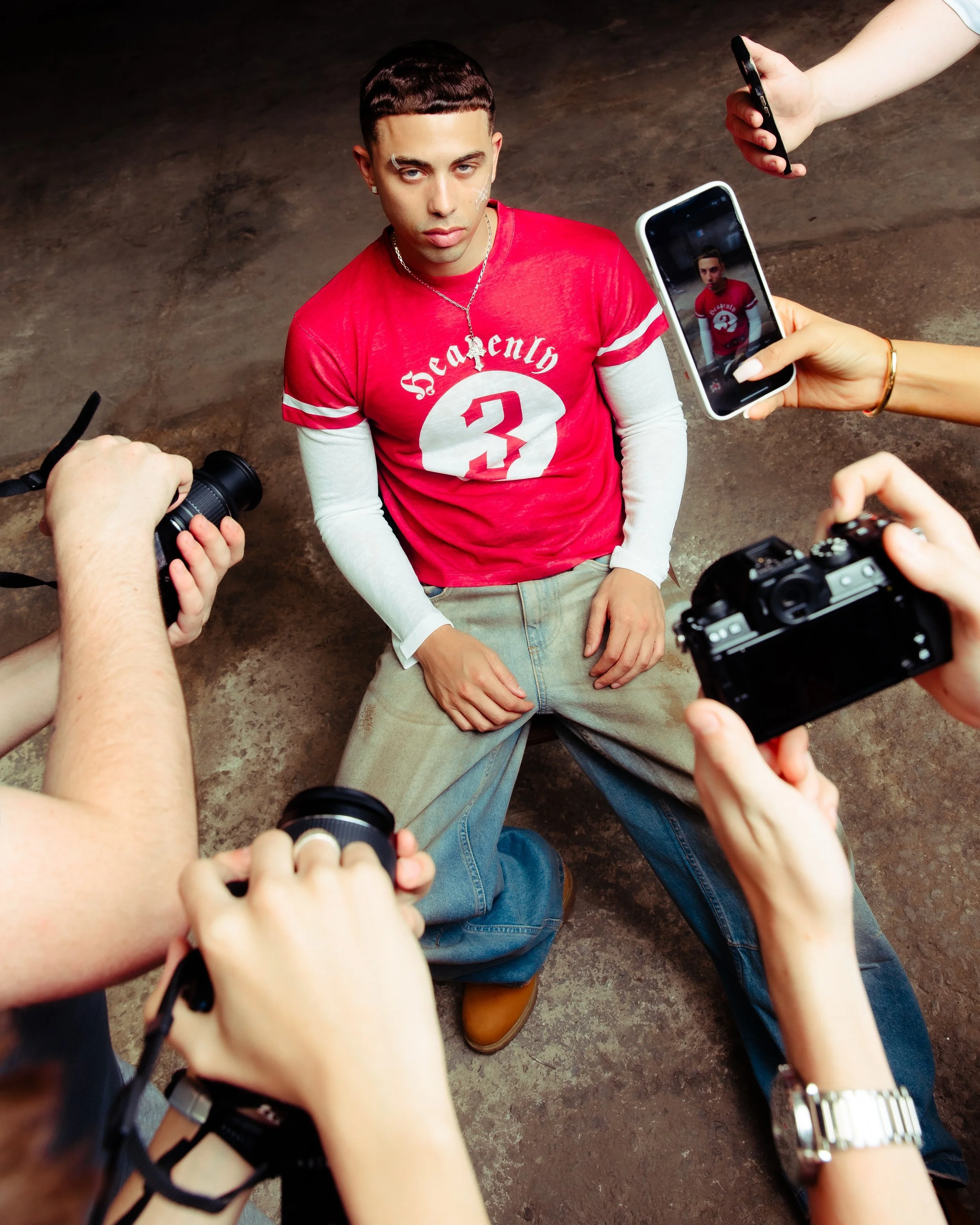 A young man with dark hair, wearing a red shirt with the number 3 and the word 'Seriously' on it, poses for photos taken by multiple people in a photo shoot. The setting appears to be indoors on a brown floor.
