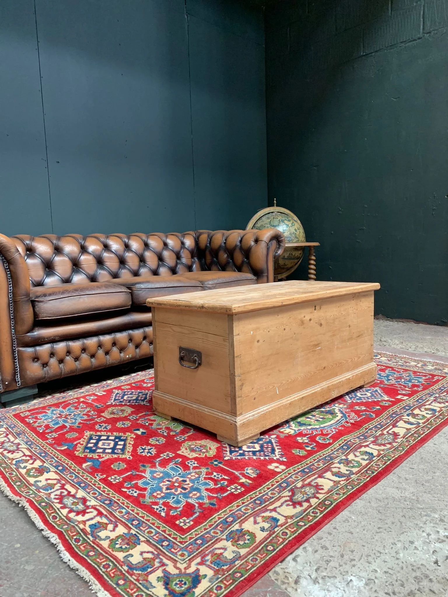 A vintage room with a brown leather Chesterfield sofa, a wooden chest/coffee table, a colorful ornate rug, and a globe on a small table in the background against dark green walls.