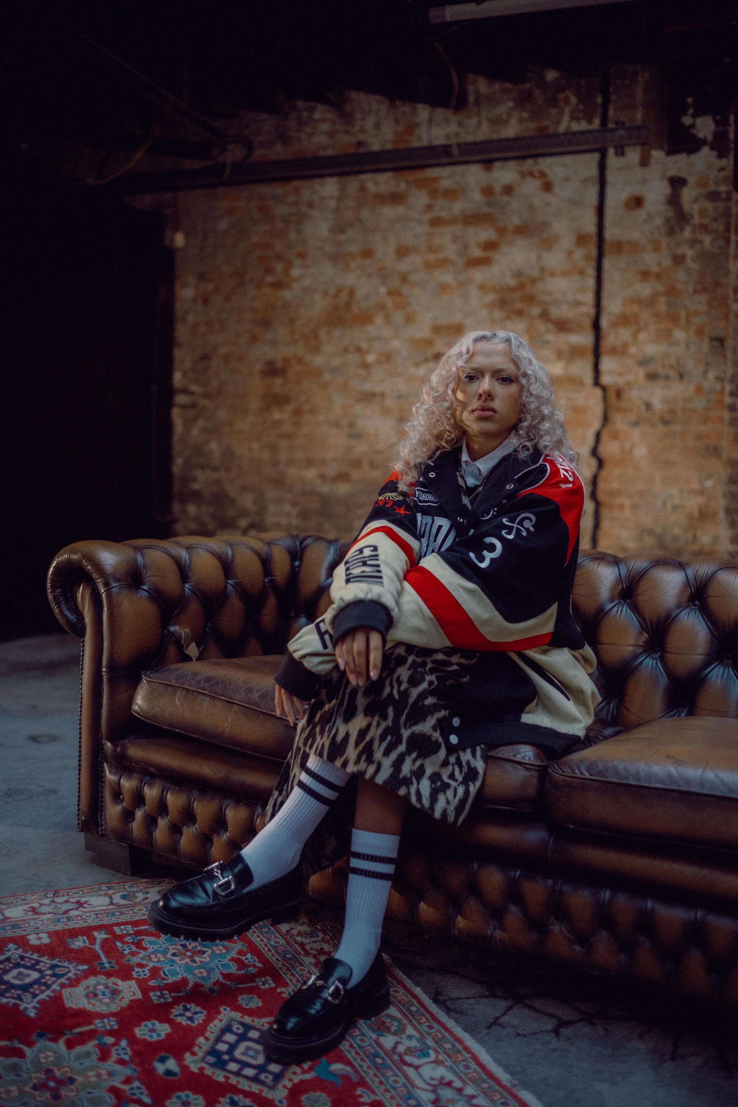 A woman with curly blonde hair sitting on a brown leather chesterfield sofa in an industrial-style room with an exposed brick wall.