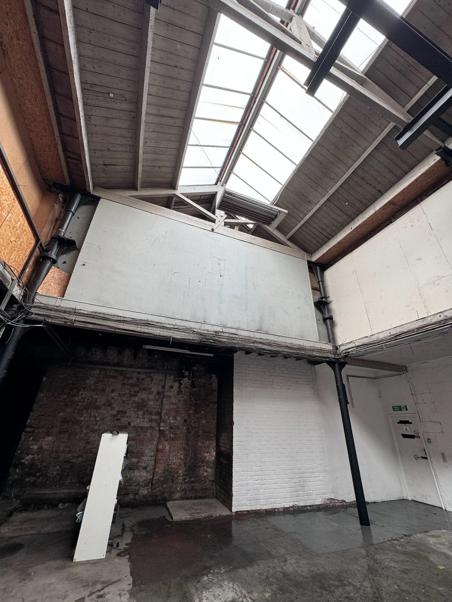 Interior of a building with exposed brick and painted white walls, a rainwater puddle on the concrete floor, and an open skylight in the wooden ceiling.