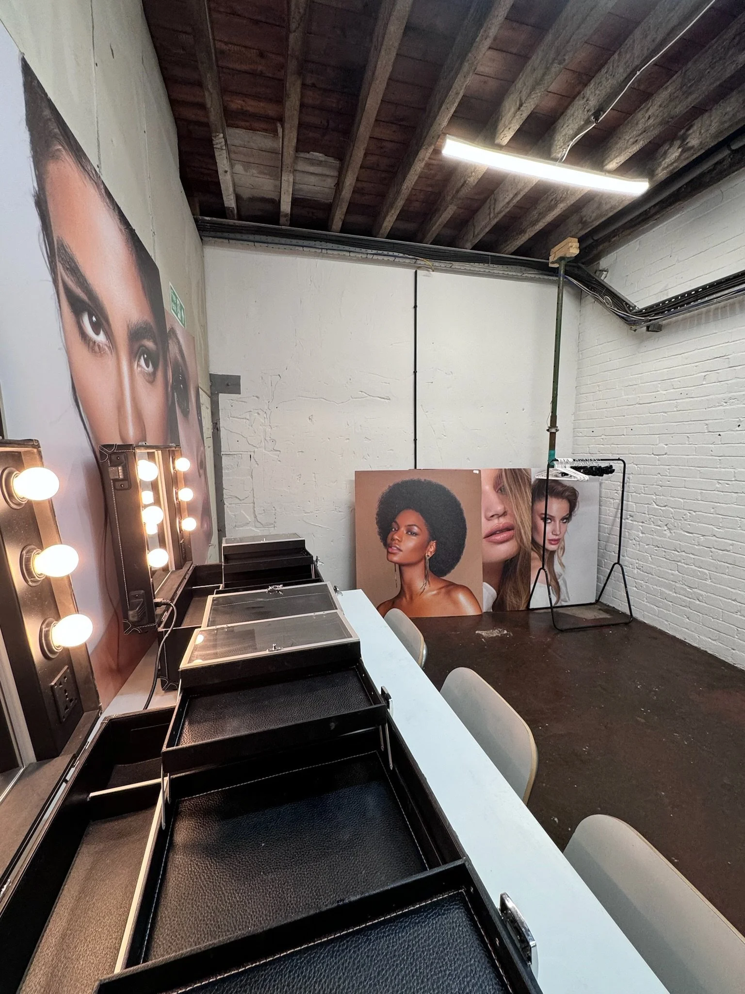A makeup station with large posters of diverse models, mirrors with lights, and empty trays, in a room with white brick walls and wooden ceiling beams.