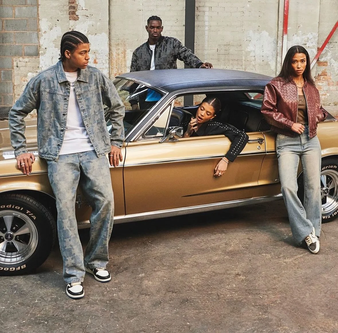 Four young adults with stylish clothing and hairstyles posing around a vintage gold and black car in an industrial setting with exposed brick and concrete walls.