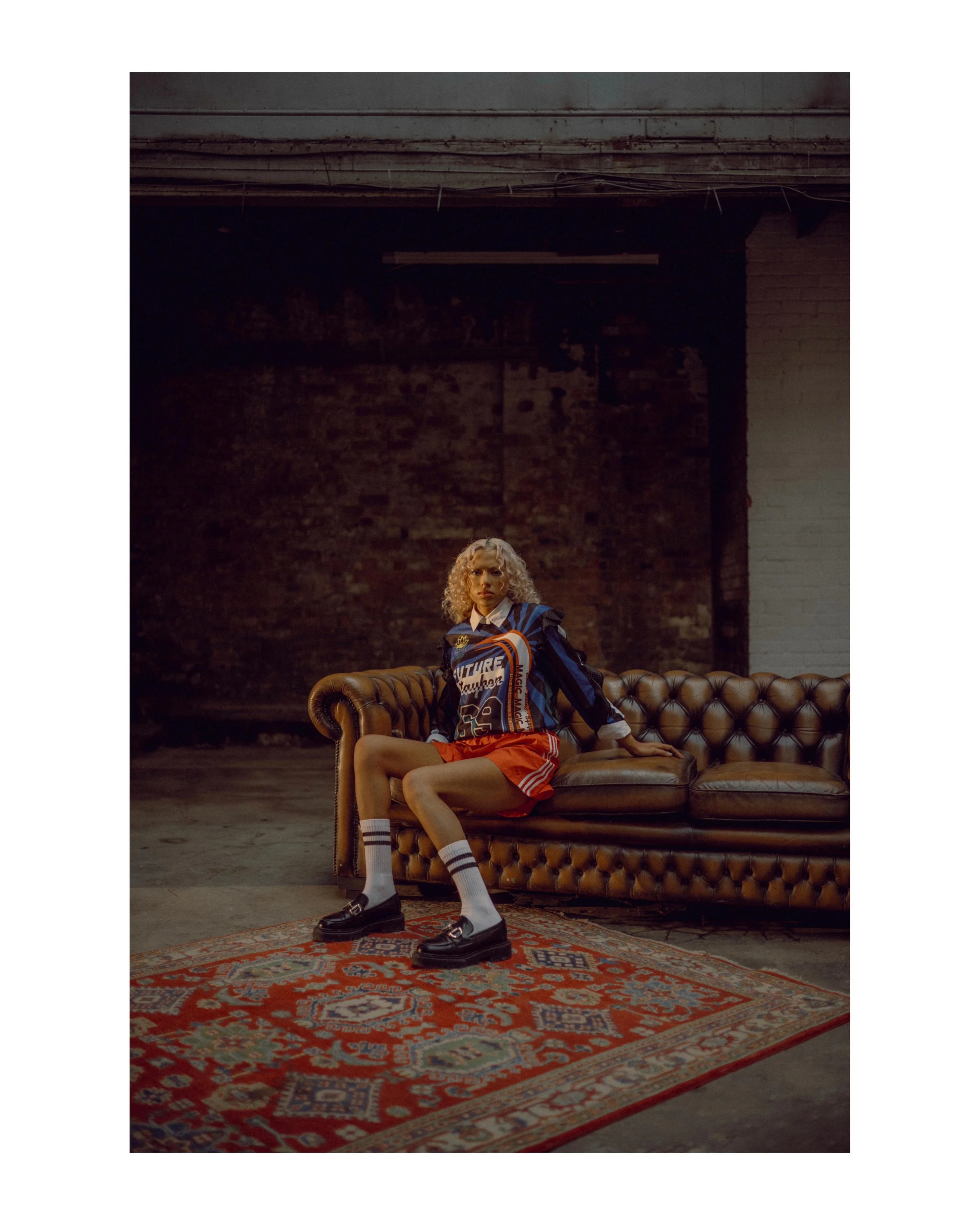 A young woman with curly blonde hair sitting on a vintage brown leather sofa in an industrial-style room with exposed brick wall and a patterned rug on the floor. She is wearing a colorful sports jacket, red shorts, white socks, and black shoes, with