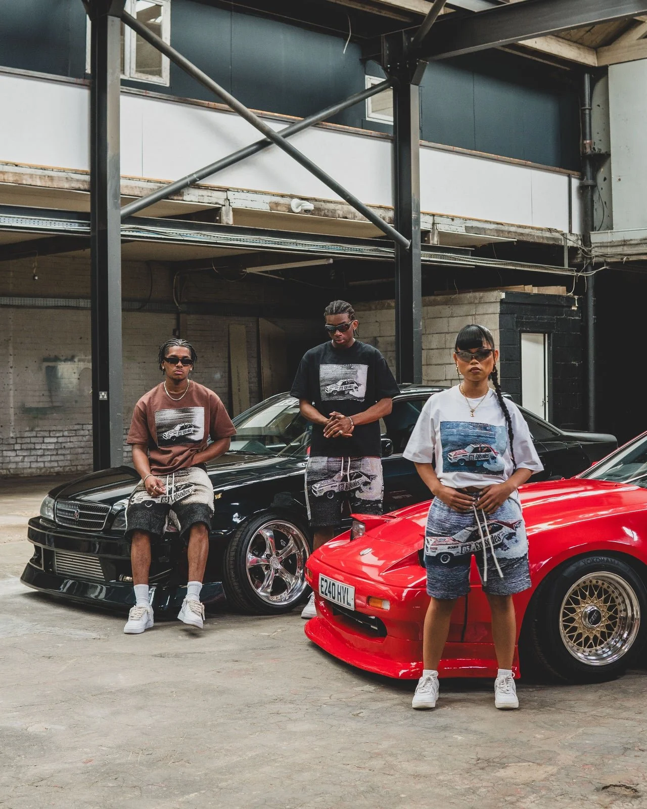 Three young people with sunglasses standing in an industrial garage with two lowrider cars, one black and one red, wearing graphic t-shirts and shorts.