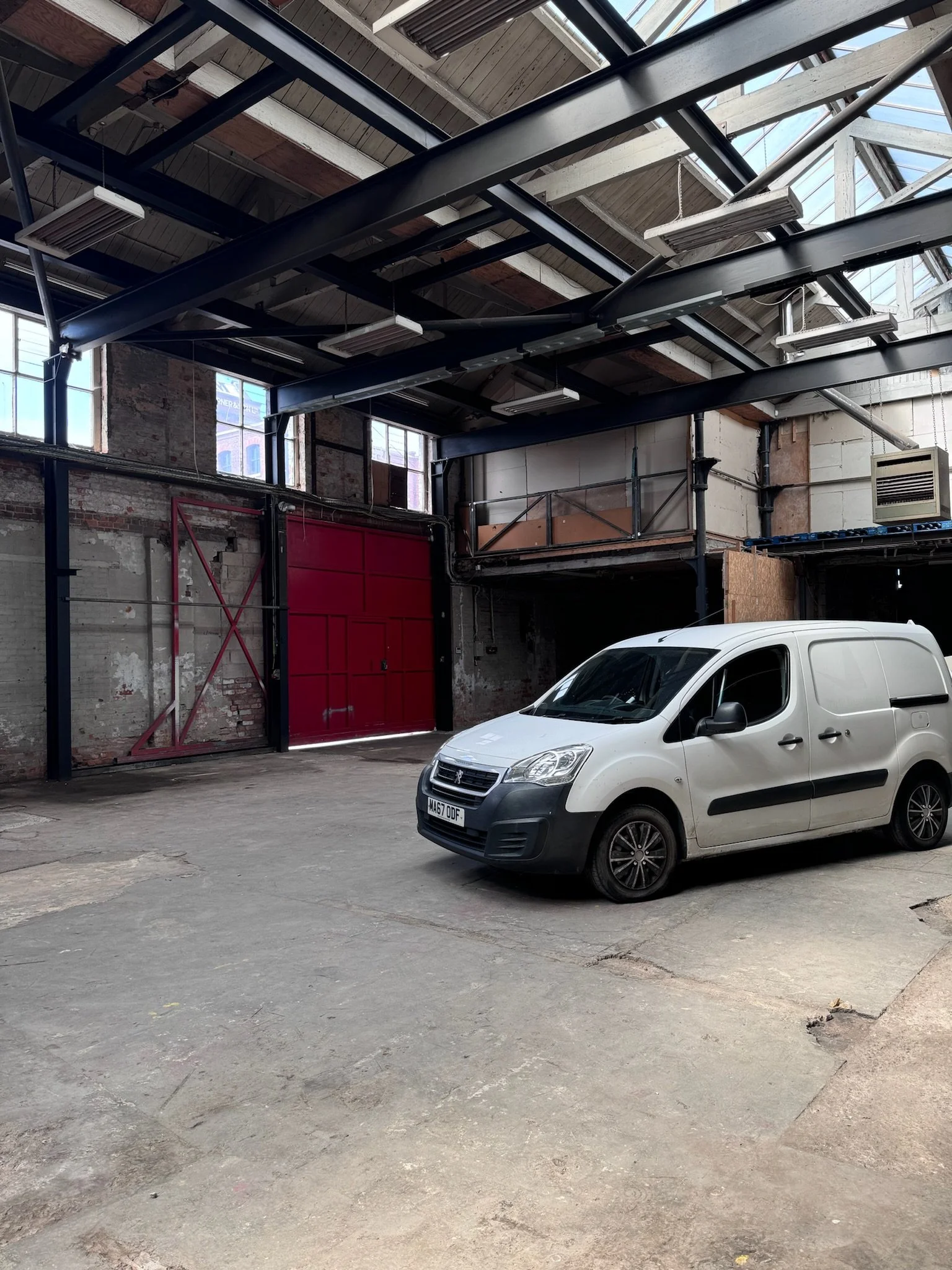 An indoor industrial space with a white van parked on a concrete floor. The space features exposed brick walls, metal beams, large windows, and a red sliding door.