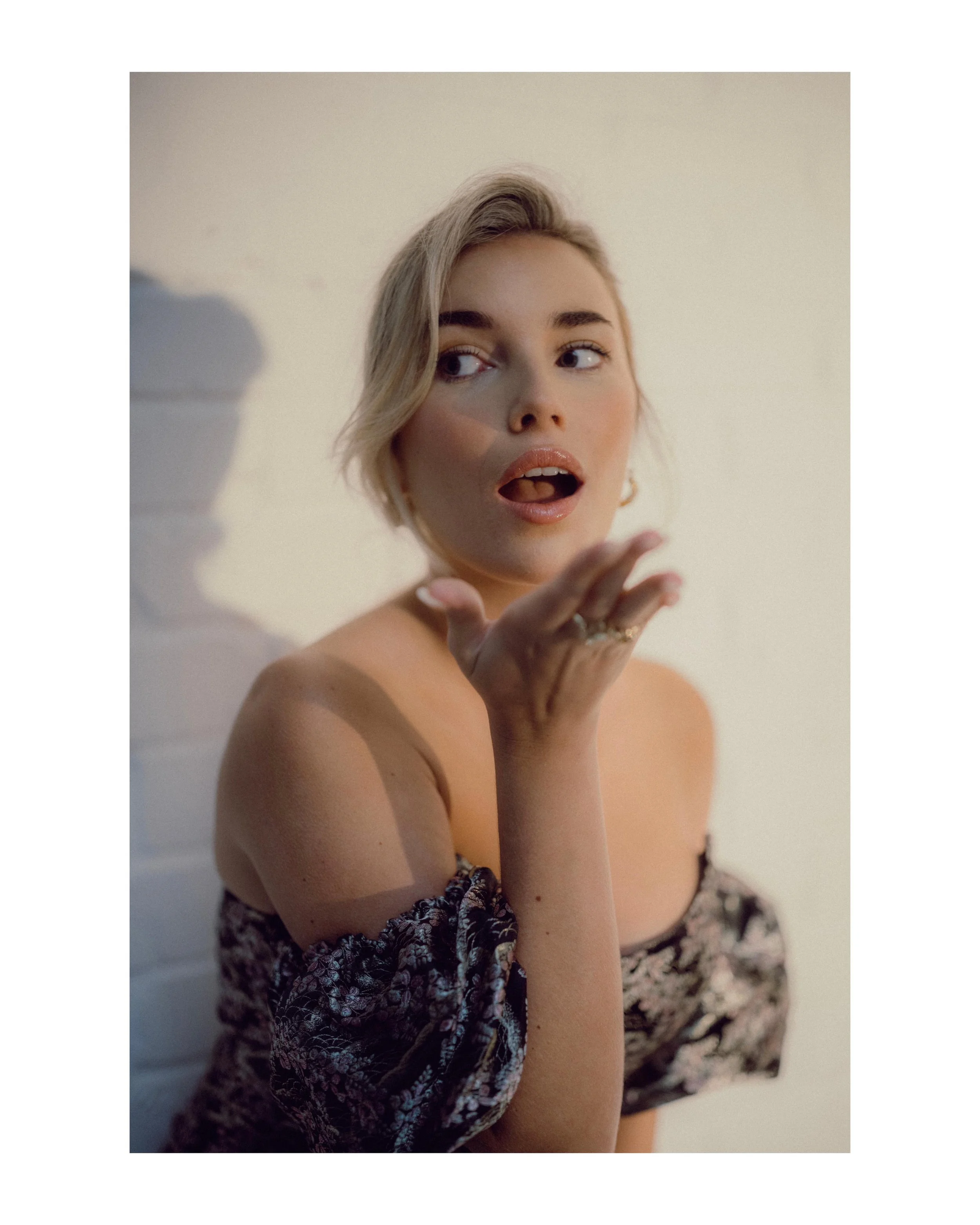 A woman with short blonde hair blowing a kiss towards the camera, wearing an off-the-shoulder floral top, with a neutral background.