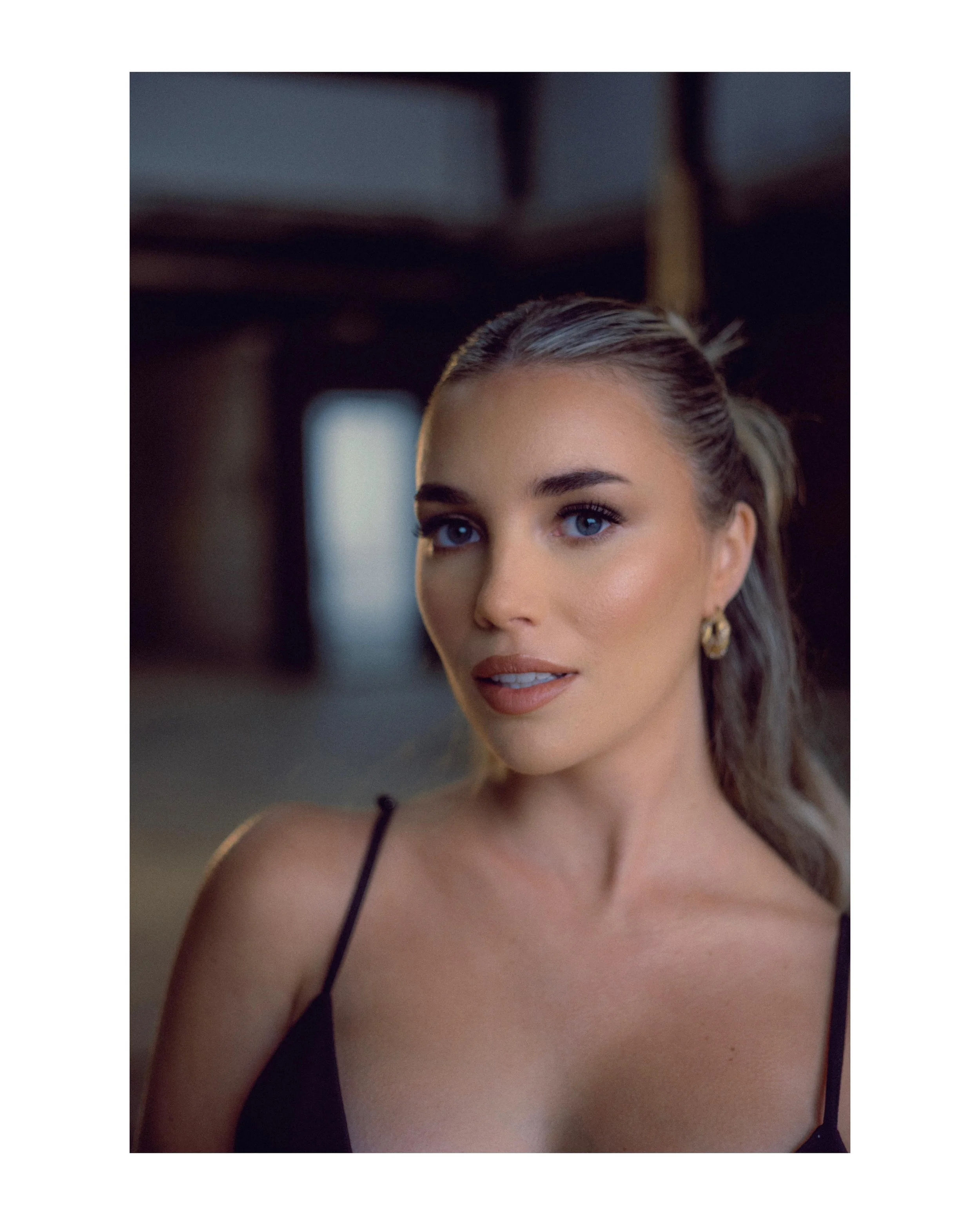 A woman with blue eyes, wearing makeup, gold earrings, and a black spaghetti strap top, looking slightly to the left inside a dimly lit building.