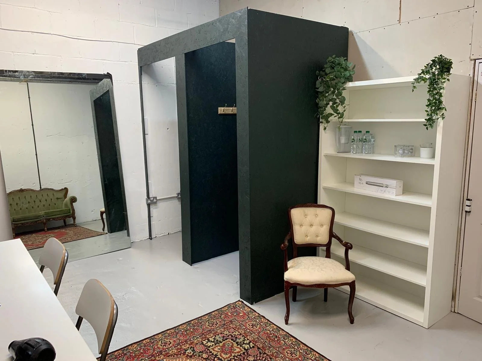 Interior of a room with a white bookshelf holding water bottles and glassware, a vintage armchair, a mirror reflecting a green sofa, and a black partition with a doorway, all on a white floor with a red patterned rug.