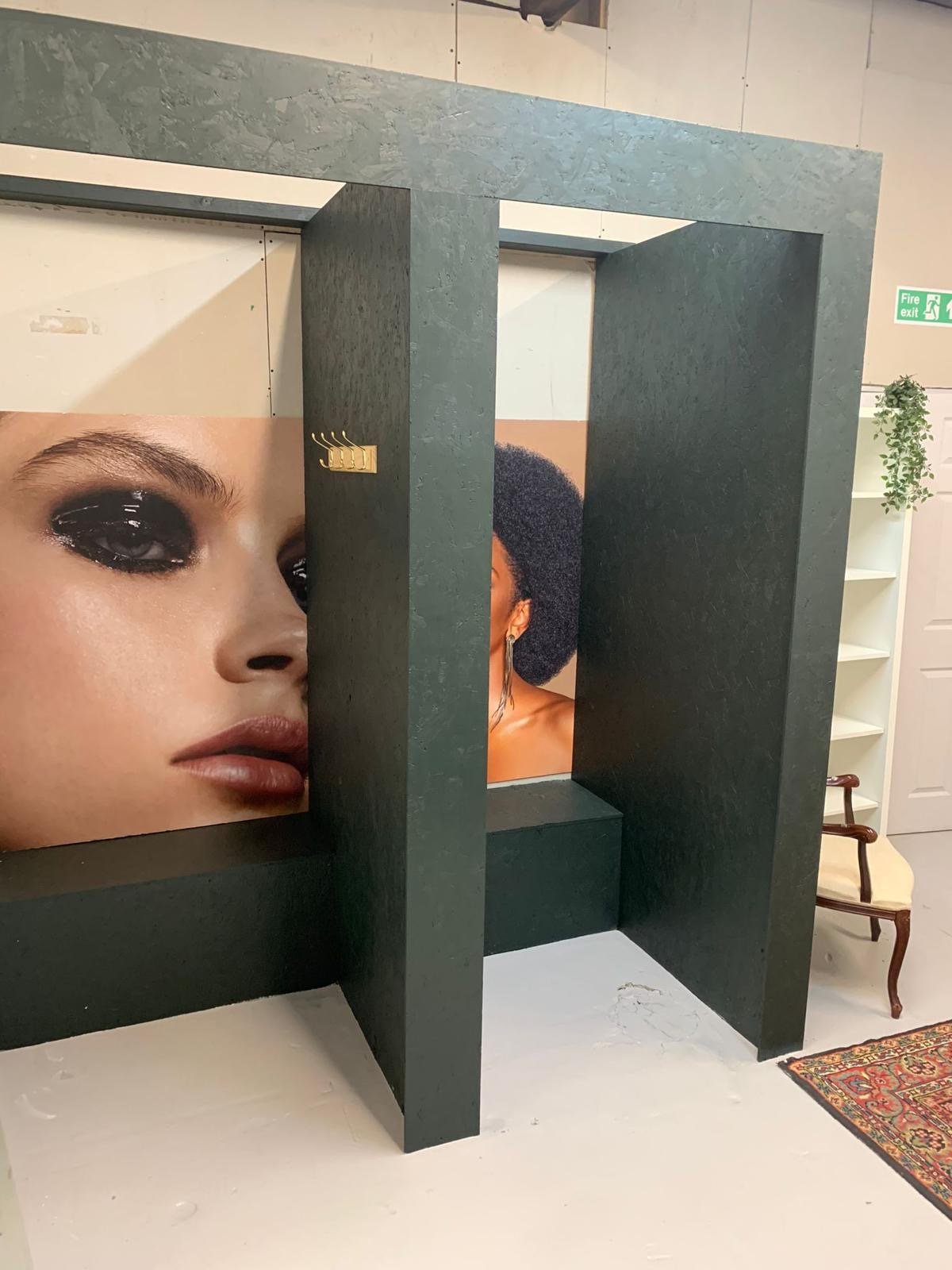 Interior of a store or gallery with posters of women, one with dark eye makeup and another with a large afro hairstyle, separated by dark green structural elements, with a sign indicating a fire exit in the background.