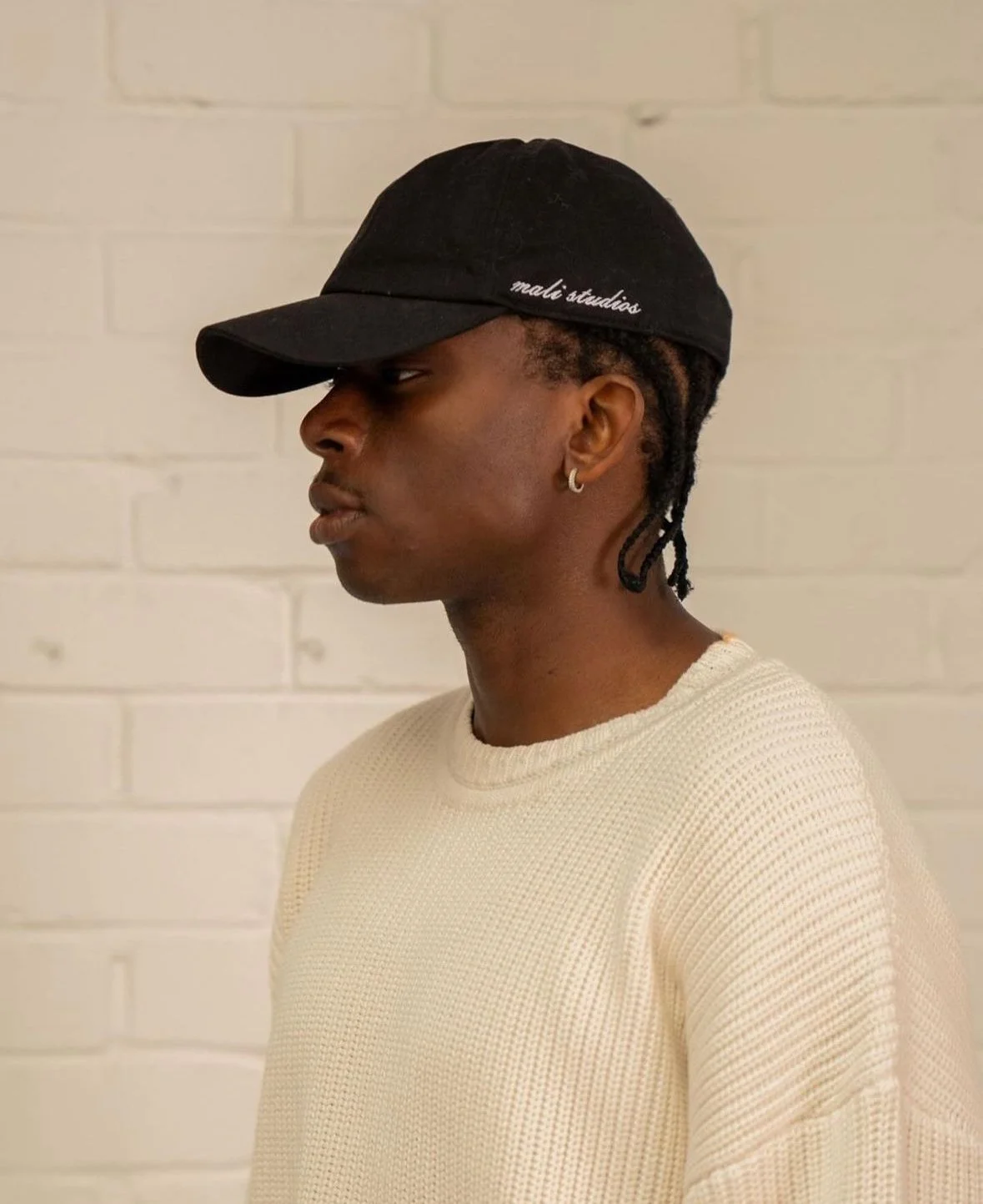 Side profile of a young man wearing a black baseball cap and a cream-colored sweater, standing against a white brick wall.
