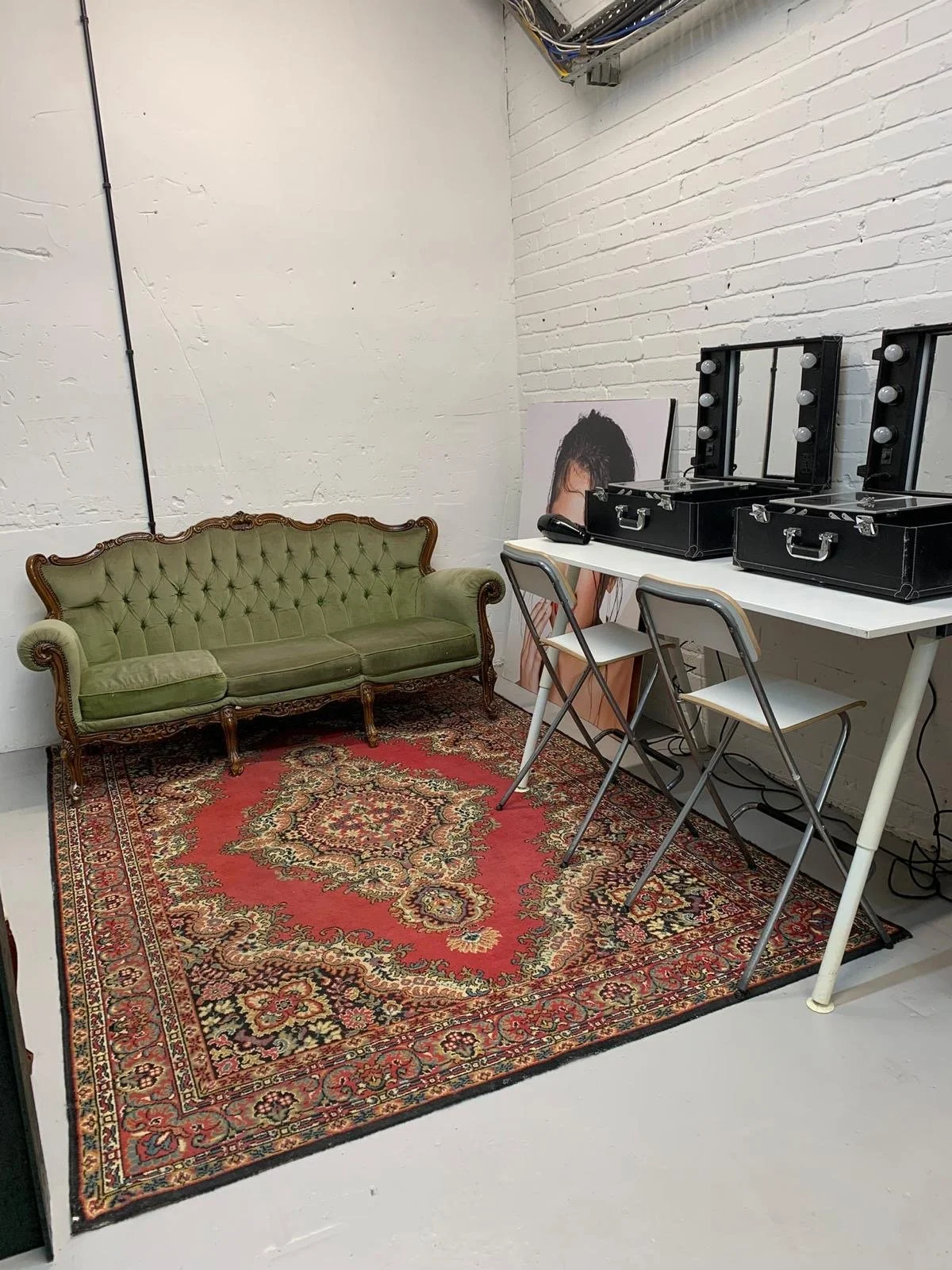 A vintage green tufted sofa on a Persian-style red rug in a room with white brick walls, a framed photo of a woman, and a white table with three lighted makeup mirrors and two chairs.