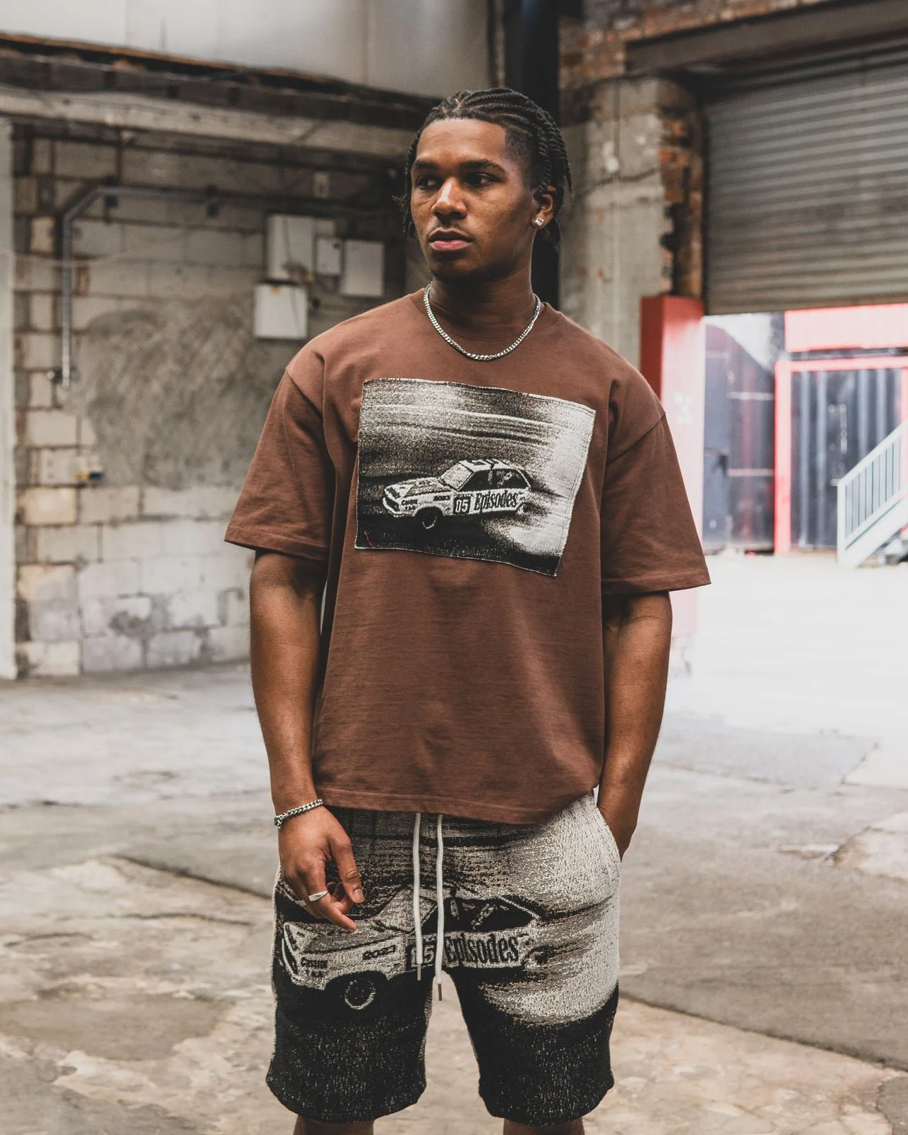 Young man standing in front of a brick wall in an industrial setting, wearing a brown T-shirt with a graphic of a car and text, and matching shorts with similar graphic design.