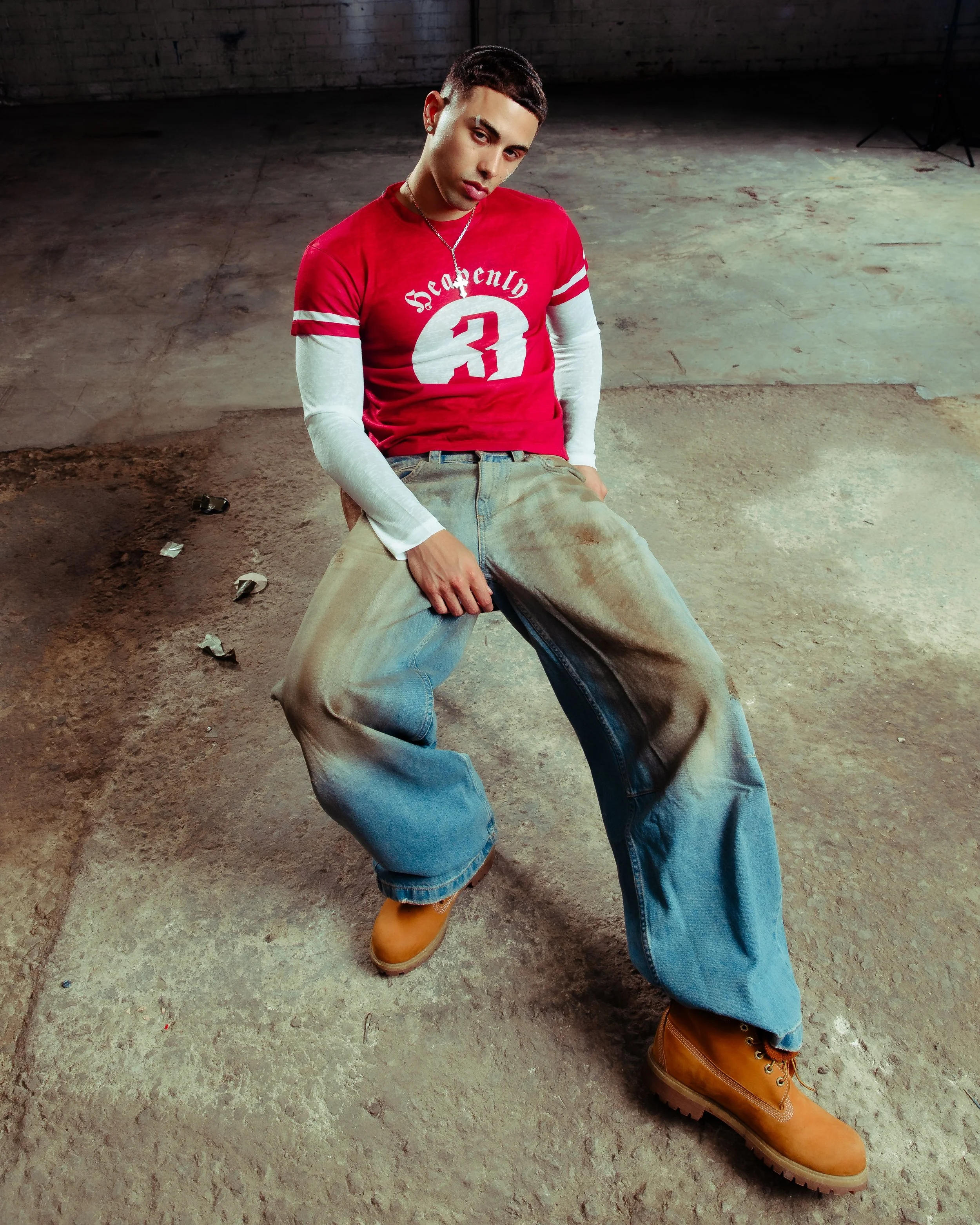 A young man is sitting on the ground in a warehouse-like space with a concrete floor and brick wall, wearing a red and white long-sleeve shirt, large baggy jeans, and tan work boots, looking at the camera with a serious expression.
