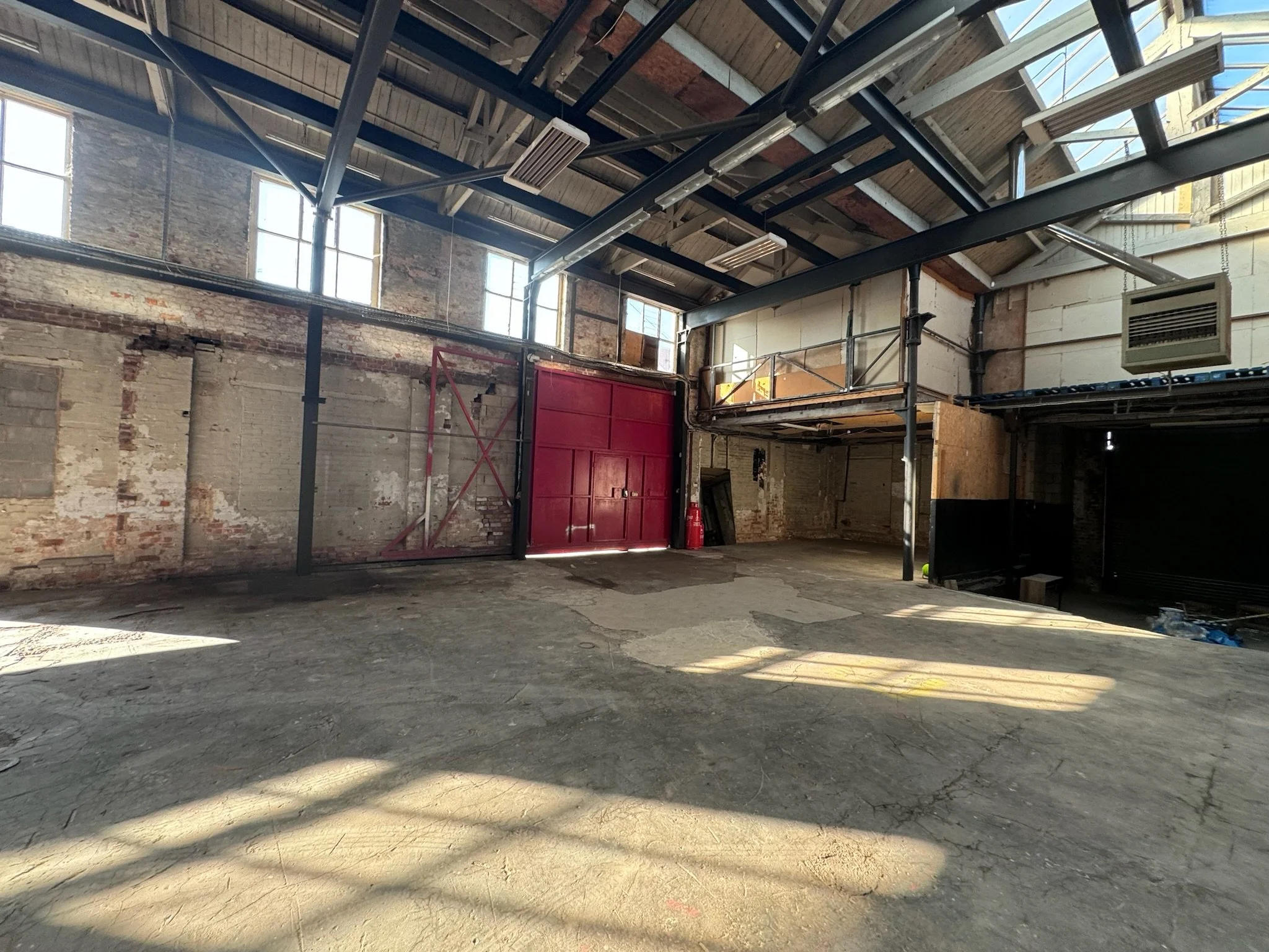 Empty industrial warehouse space with exposed brick walls, large windows, a red sliding door, and a mezzanine level with metal railing.