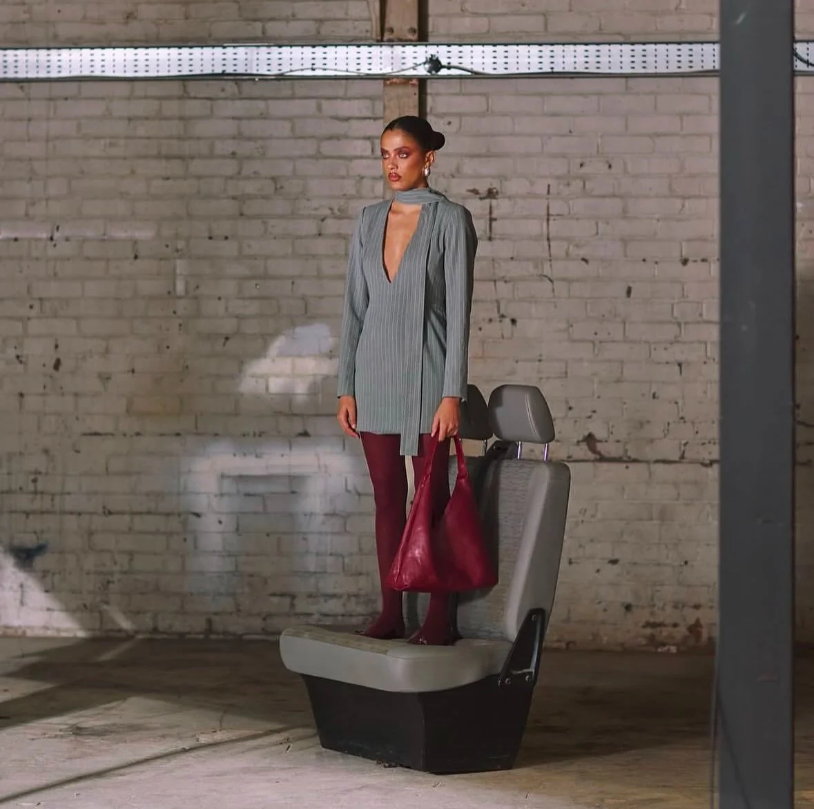 A woman in a gray blazer-style dress stands on a car seat in front of a brick wall, holding a large maroon handbag. She is wearing maroon tights and styled hair, with earrings.
