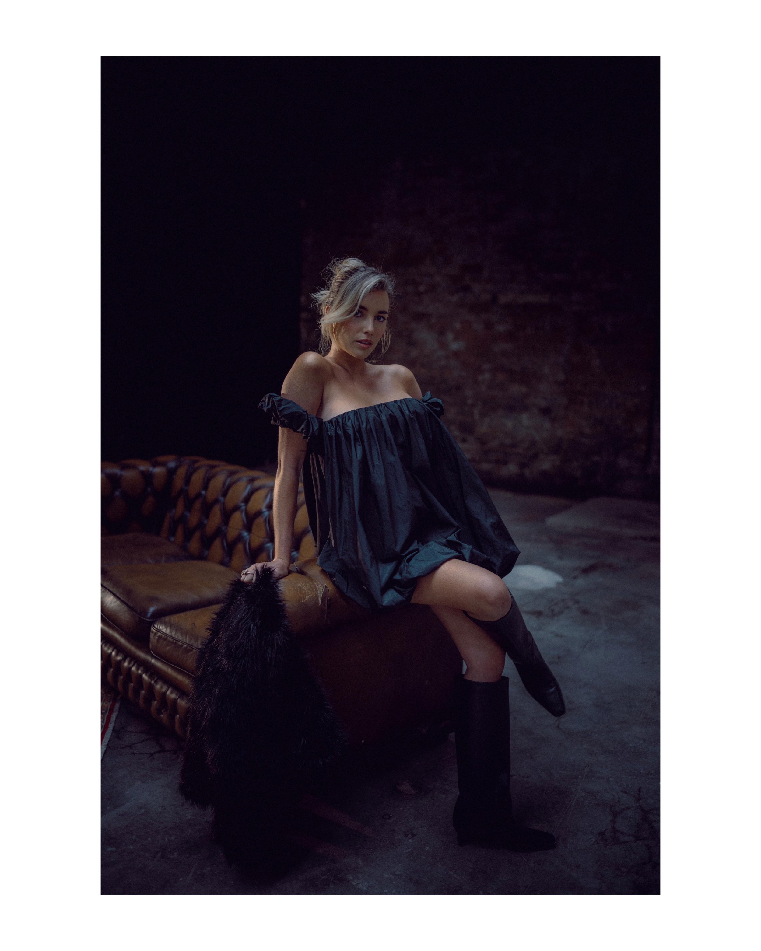 A woman in a dark off-the-shoulder dress sitting on a brown leather sofa in a dimly lit room with a brick wall background, holding a black fur handbag and wearing knee-high black boots.