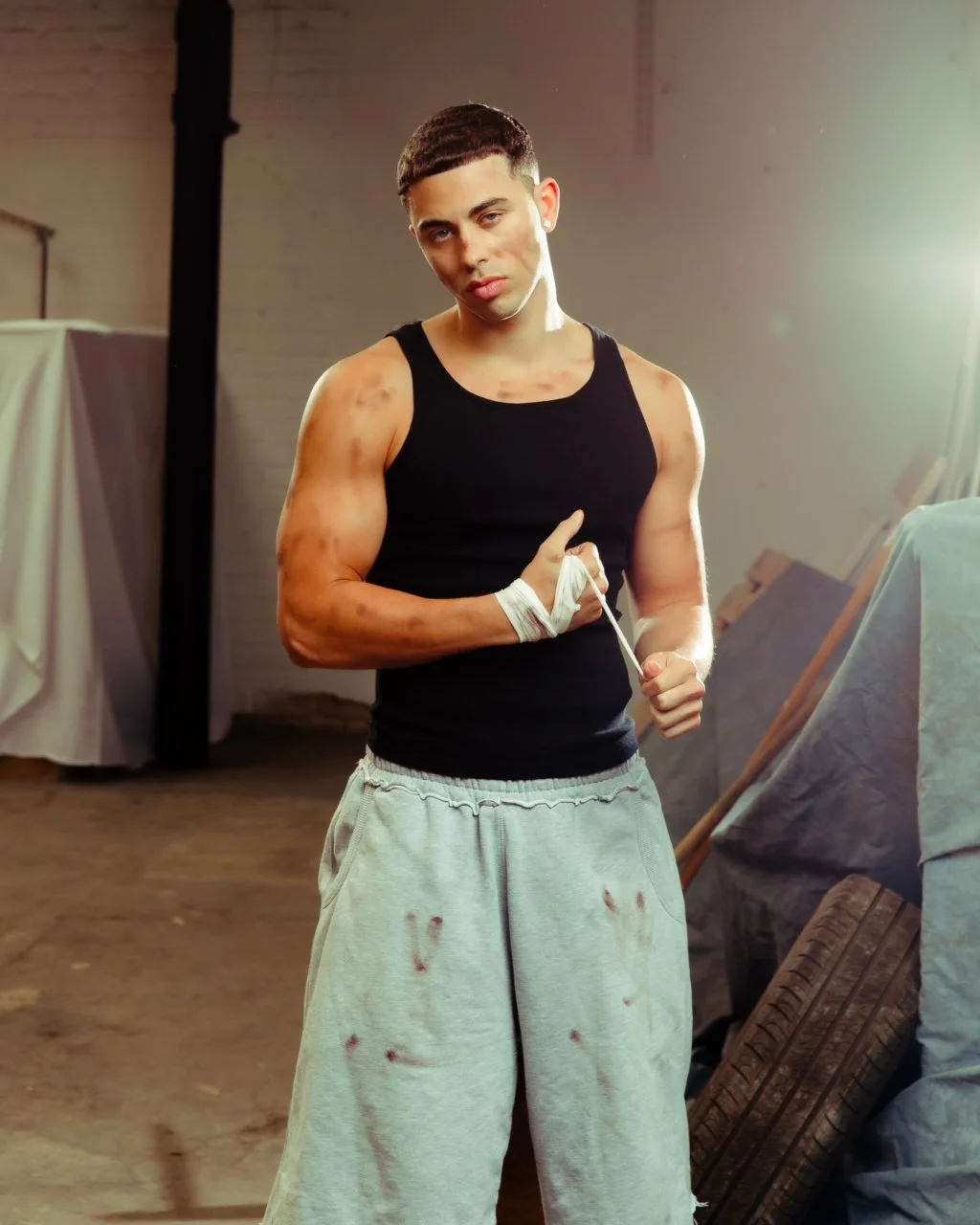 Young man with bruises and cuts on his arms and face, wearing a black tank top and gray sweatpants stained with dirt, standing in a dimly lit industrial space.