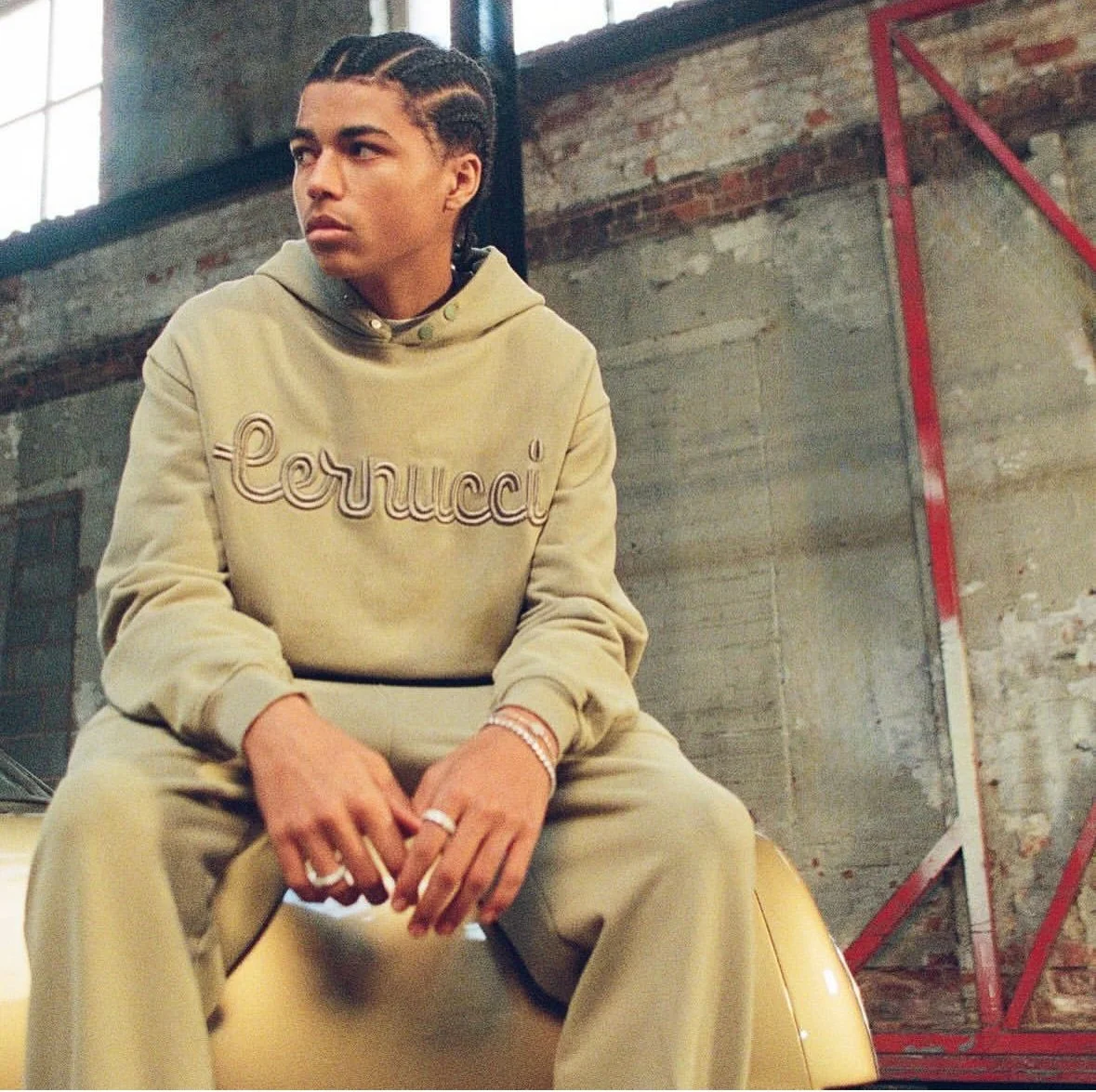 A young person with braided hair wearing a beige hoodie with 'Gucci' embroidered on it, sitting on a yellow object in an industrial-style space with exposed brick and concrete walls.