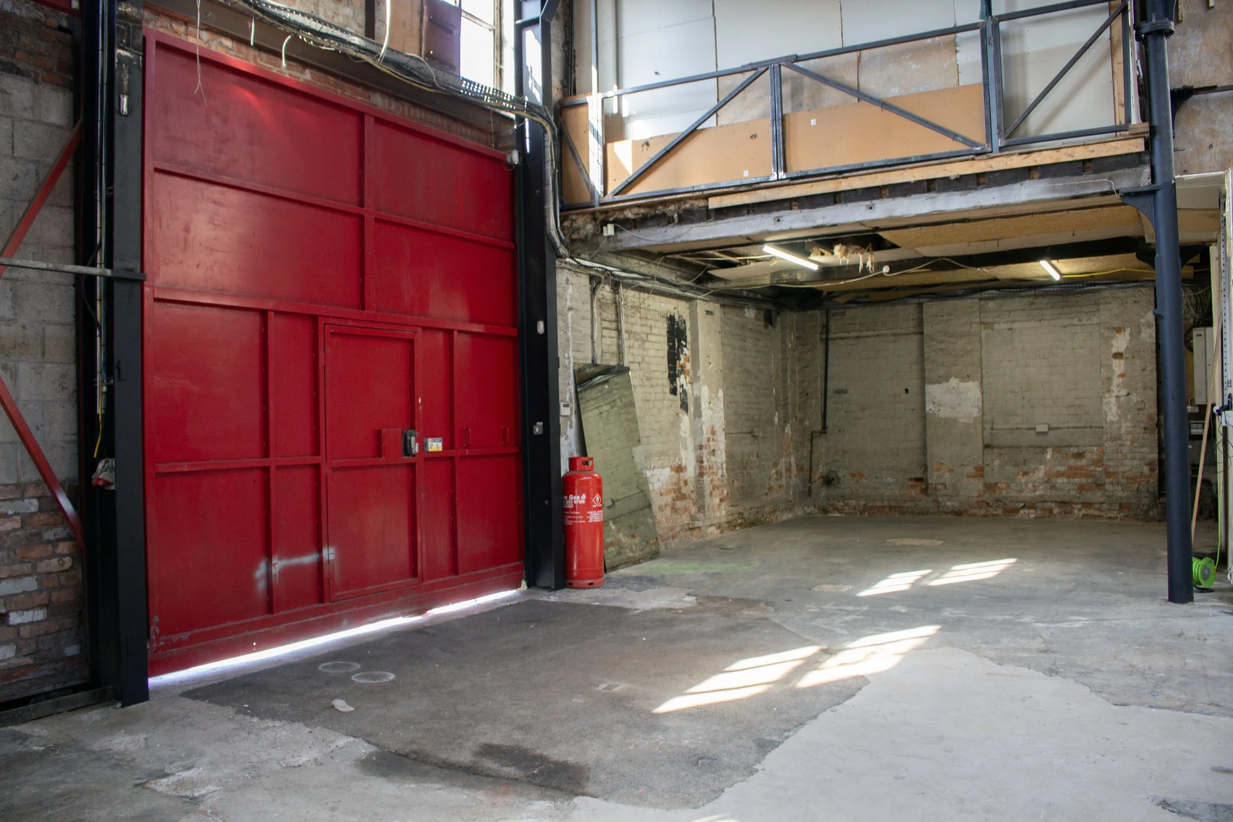An empty industrial garage with a red roll-up door, exposed brick walls, and a concrete floor, illuminated by sunlight through windows.