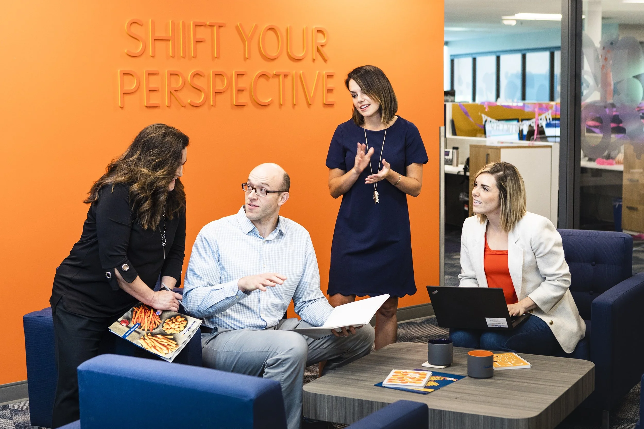 Four colleagues having a meeting in an office lounge area, with an orange wall that has the words 'Shift Your Perspective' in 3D letters.