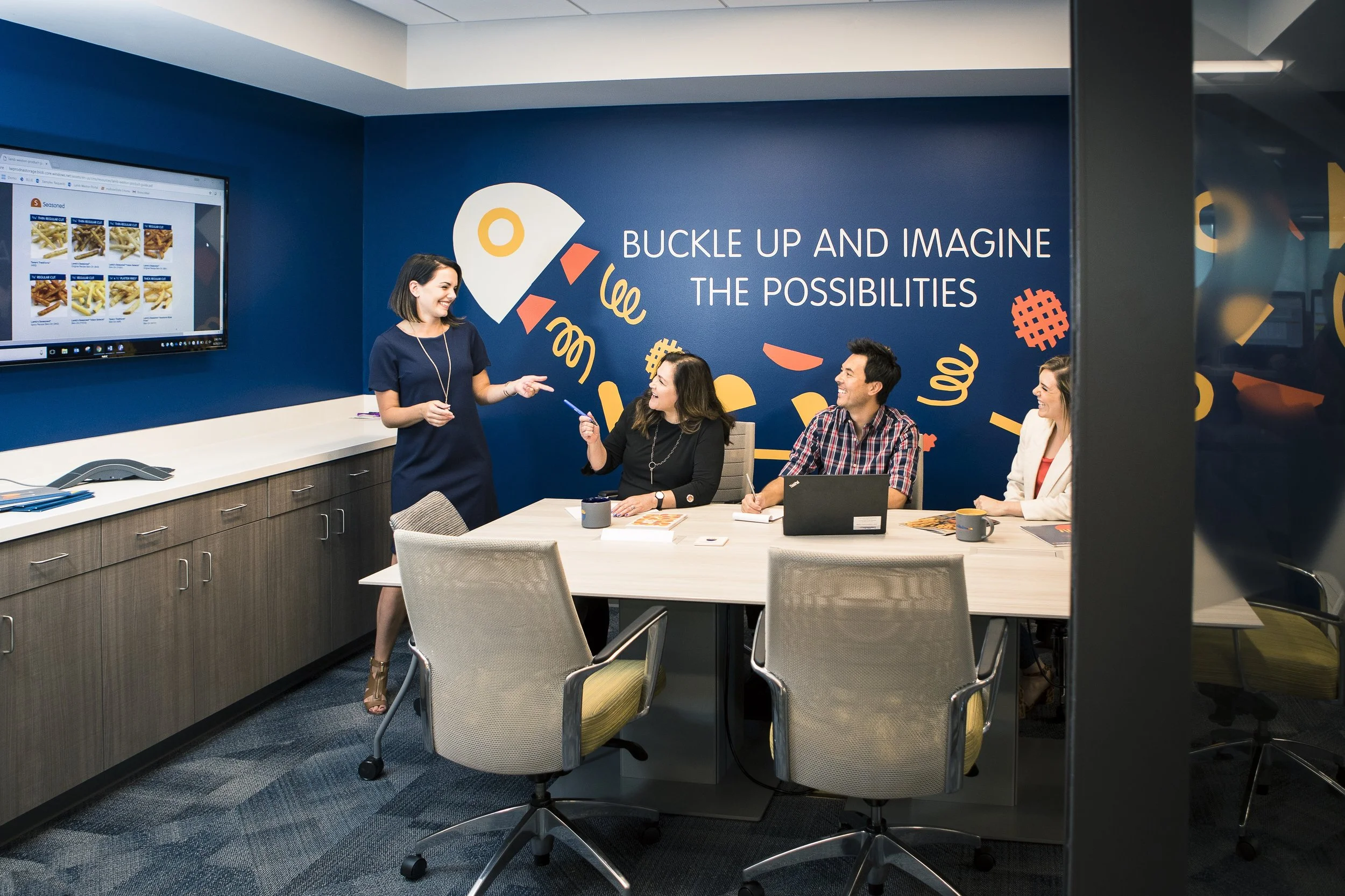 Four coworkers in a meeting room, one woman standing and smiling, three others seated at a table with laptops and notebooks, a blue wall with the words 'Buckle up and imagine the possibilities,' and colorful abstract shapes, with a large screen displaying a website.