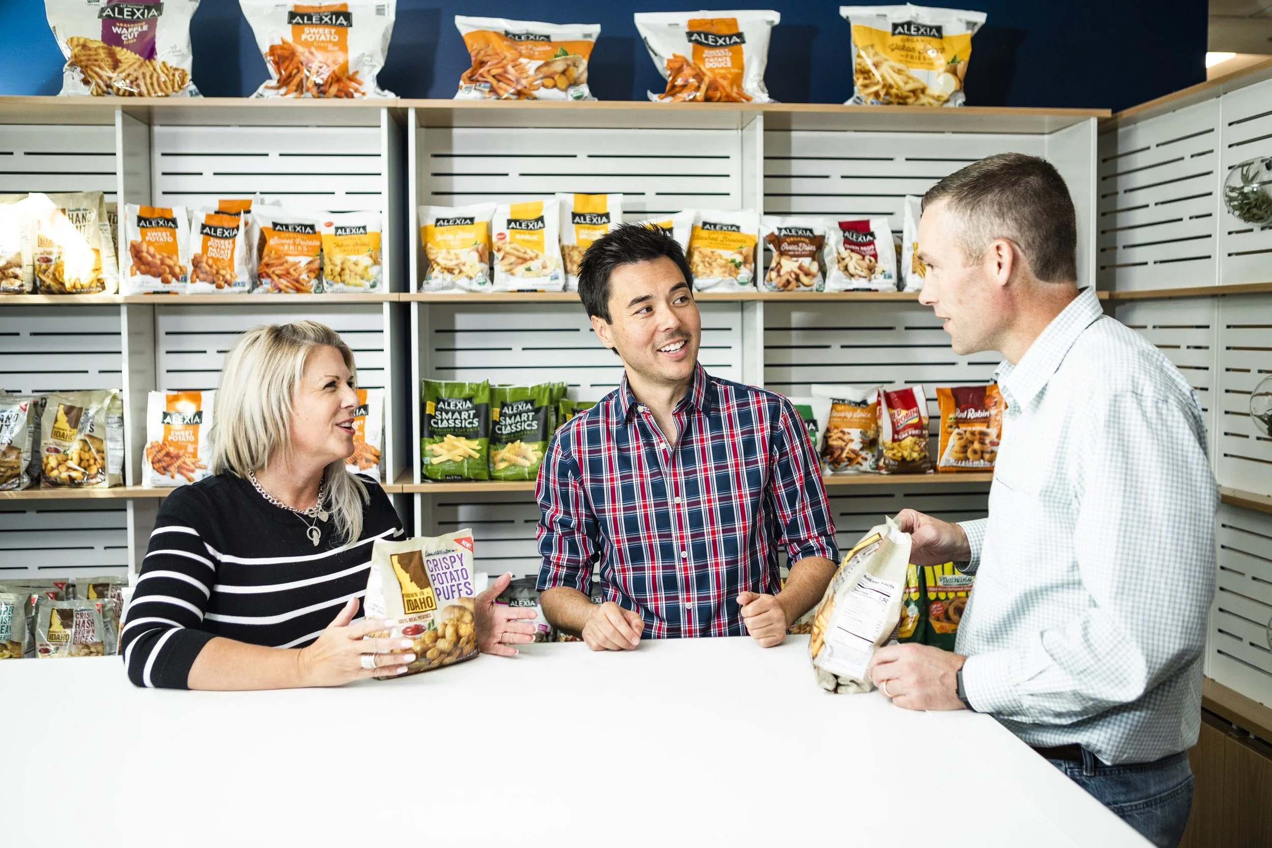 Three people in a grocery store, a woman and two men, are talking at a counter with snack bags on shelves behind them. The woman is holding a bag of crispy potato puffs, and the man on the right is holding a bag of chips, while the man in the center is smiling and engaging in conversation.