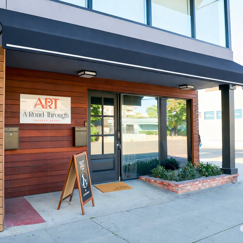 The entrance of a therapy group clinic called Art, A Road Through. The building has wood siding, a black door, two mailboxes, and a sidewalk with a small garden bed. There is a sandwich board sign outside the door with a logo of a cat and dog, indicating a pet-friendly policy.
