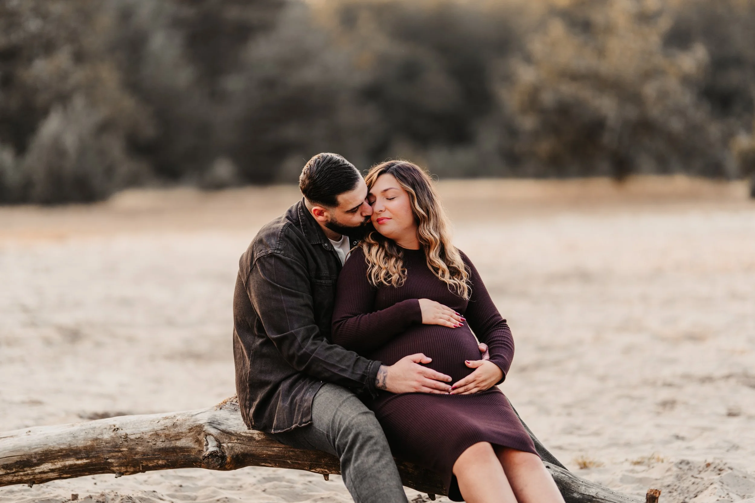 Ein schwangeres Paar sitzt auf einem Baumstamm in einer natürlichen, offenen Umgebung, umarmt sich und zeigt Zuneigung.